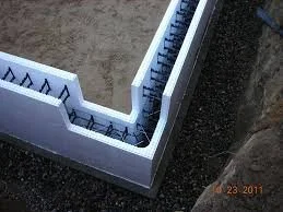 Close-up of a construction site showing the foundation walls with rebar reinforcement, surrounded by dirt and gravel.