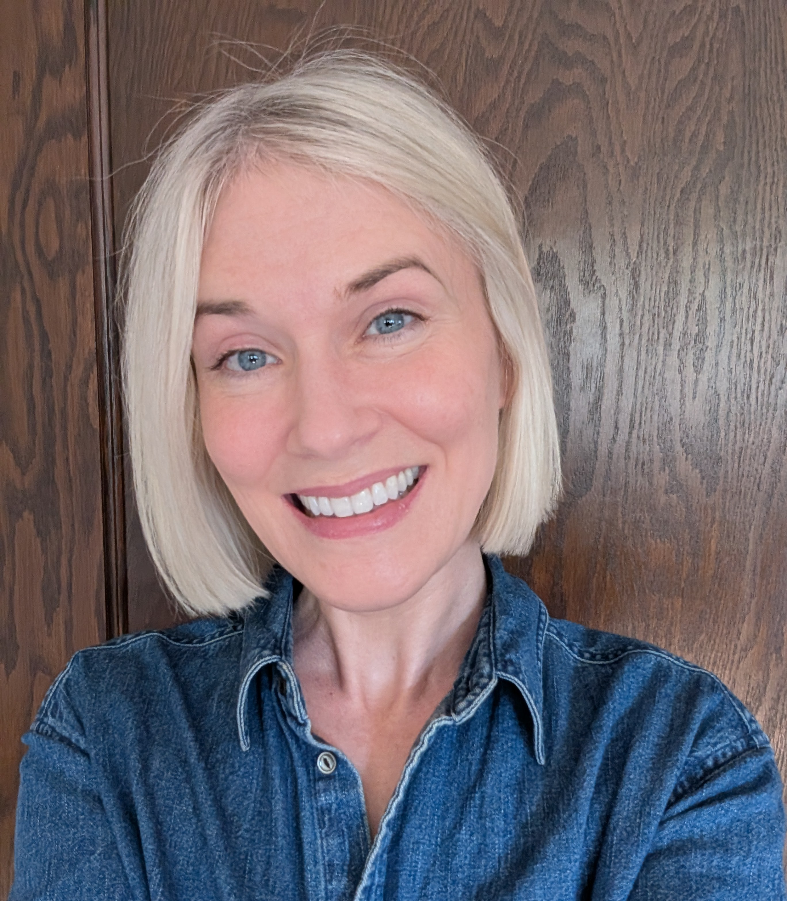 A smiling woman with blonde hair and blue eyes taking a close-up selfie in front of a wooden background, wearing a denim shirt.