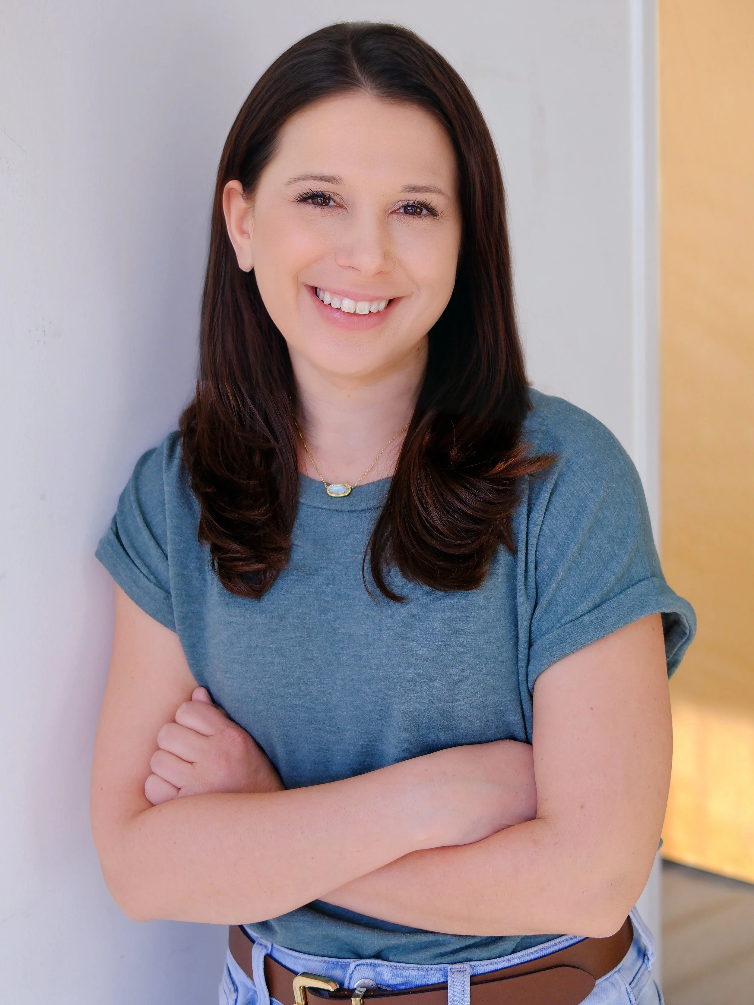 A smiling woman with dark brown hair, wearing a blue t-shirt, standing with arms crossed against a light-colored wall.