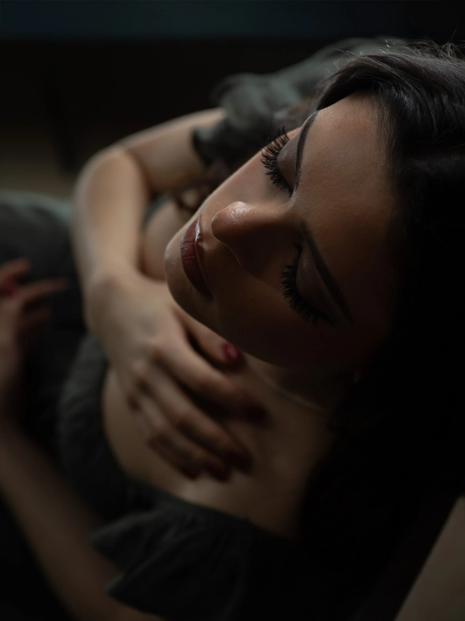 Close-up of a woman with long dark hair and makeup, lying down with eyes closed, in a dimly lit environment.