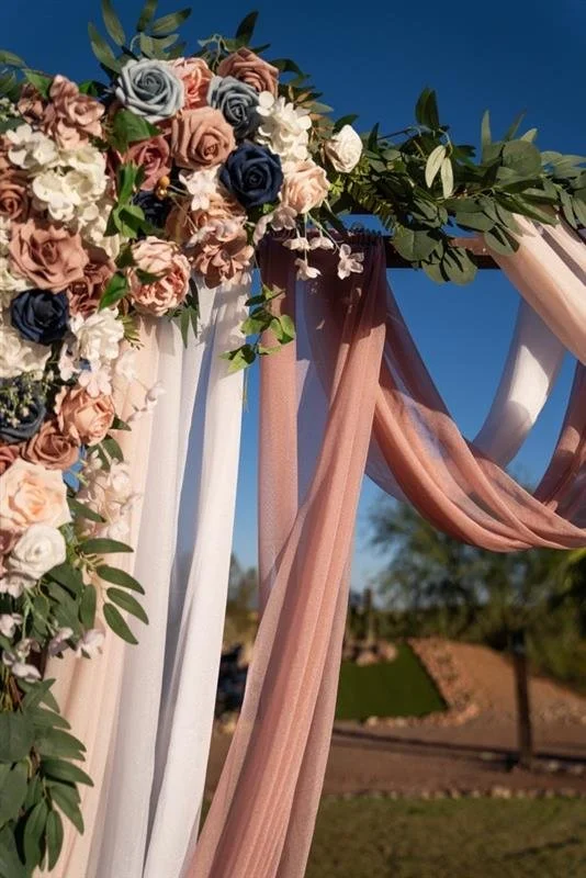 Colorful flower arrangement with pink, white, peach, gray, and navy roses intertwined with green leaves and draped pink, blush, and white fabric outdoors on a sunny day.