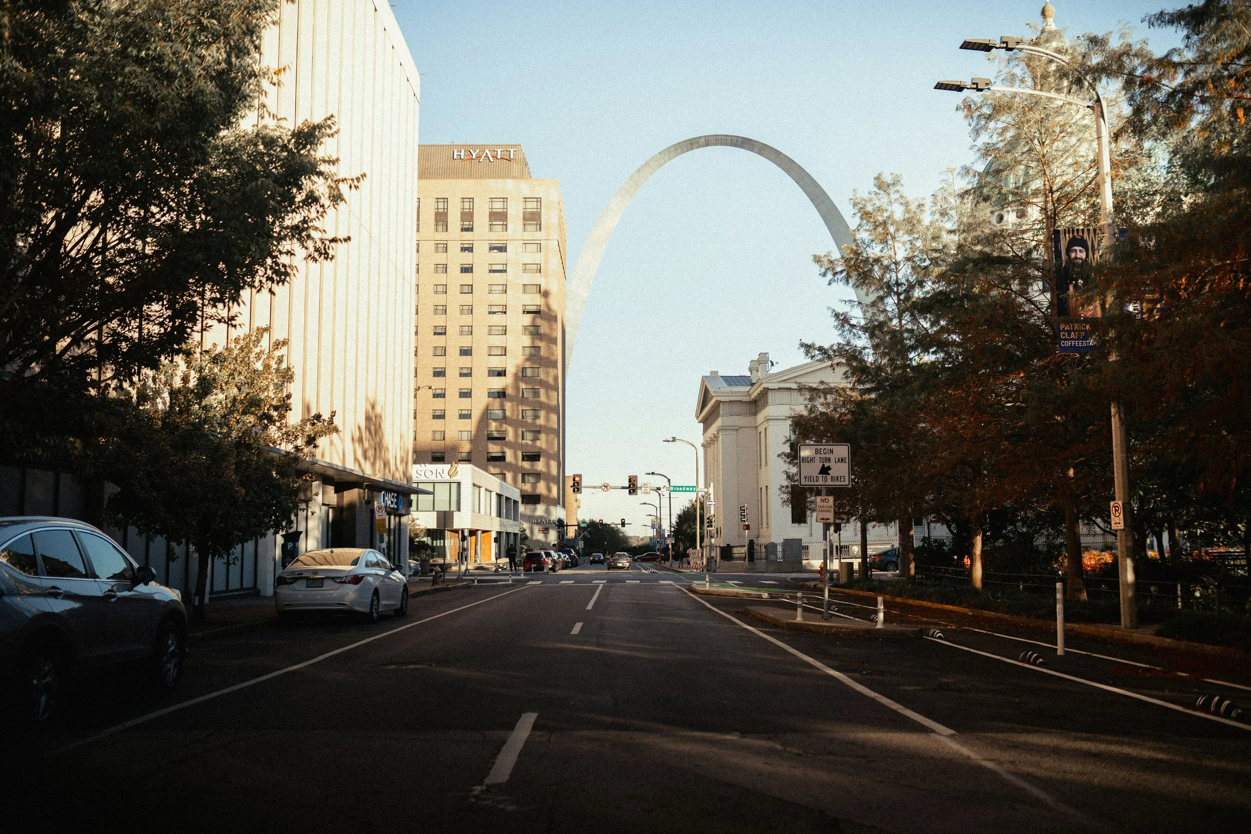Downtown street scene with cars parked on the side, trees lining the street, and the Gateway Arch visible in the background during the daytime.