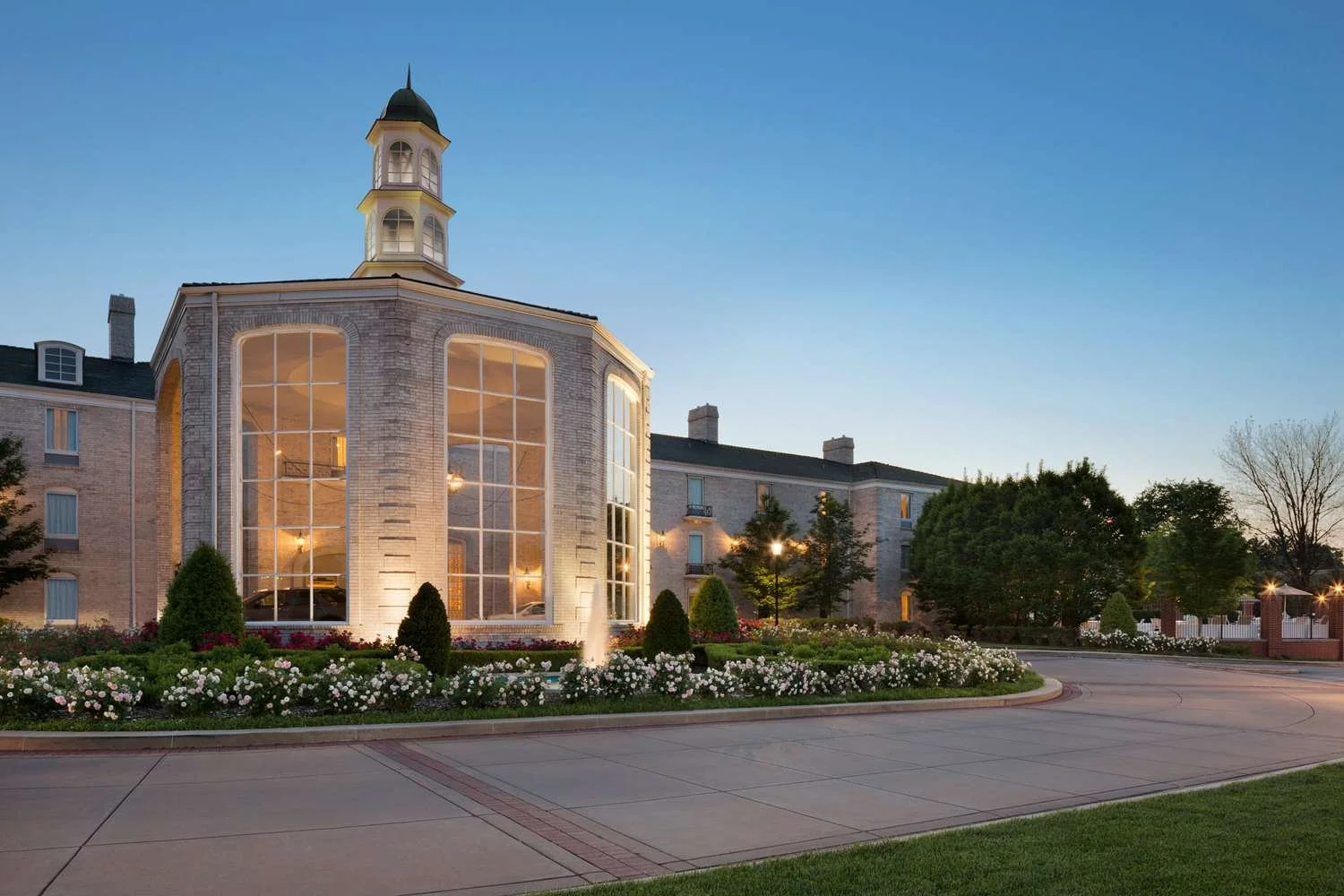 A large, elegant building with a prominent tower featuring windows and a green spire; the building has large glass windows illuminated from within. The surrounding area includes a landscaped garden with blooming flowers, trees, and a fountain, all set against a clear evening sky.
