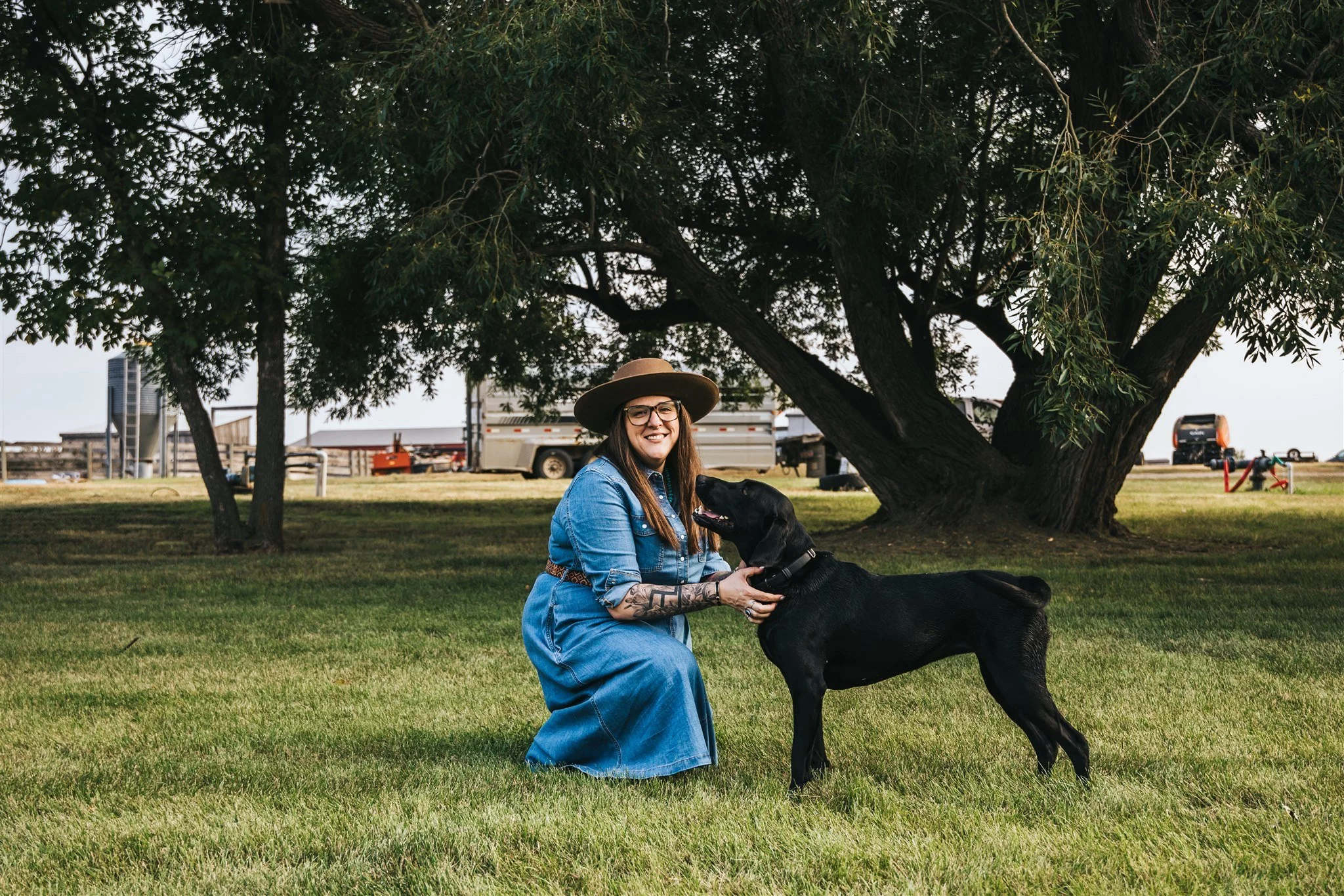 A woman wearing a blue denim dress and a wide-brimmed hat kneeling on grass and smiling at a black dog under a large tree in a park.