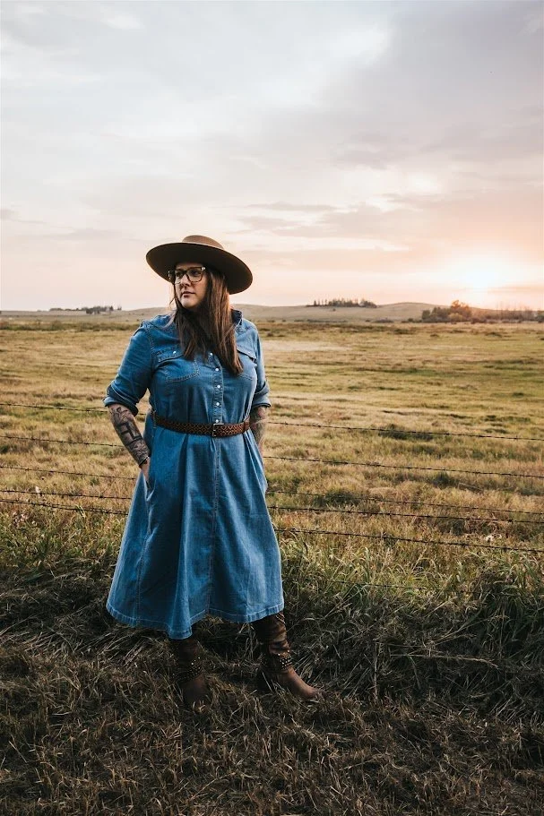 A woman with tattoos wearing glasses, a denim dress, and a wide-brimmed hat, standing in a grassy field during sunset, with a wire fence in the foreground.