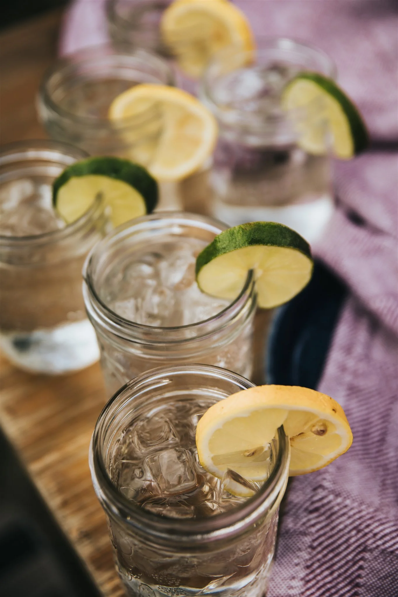 Four glasses of iced water with lemon and lime slices on a wooden surface with a purple cloth.