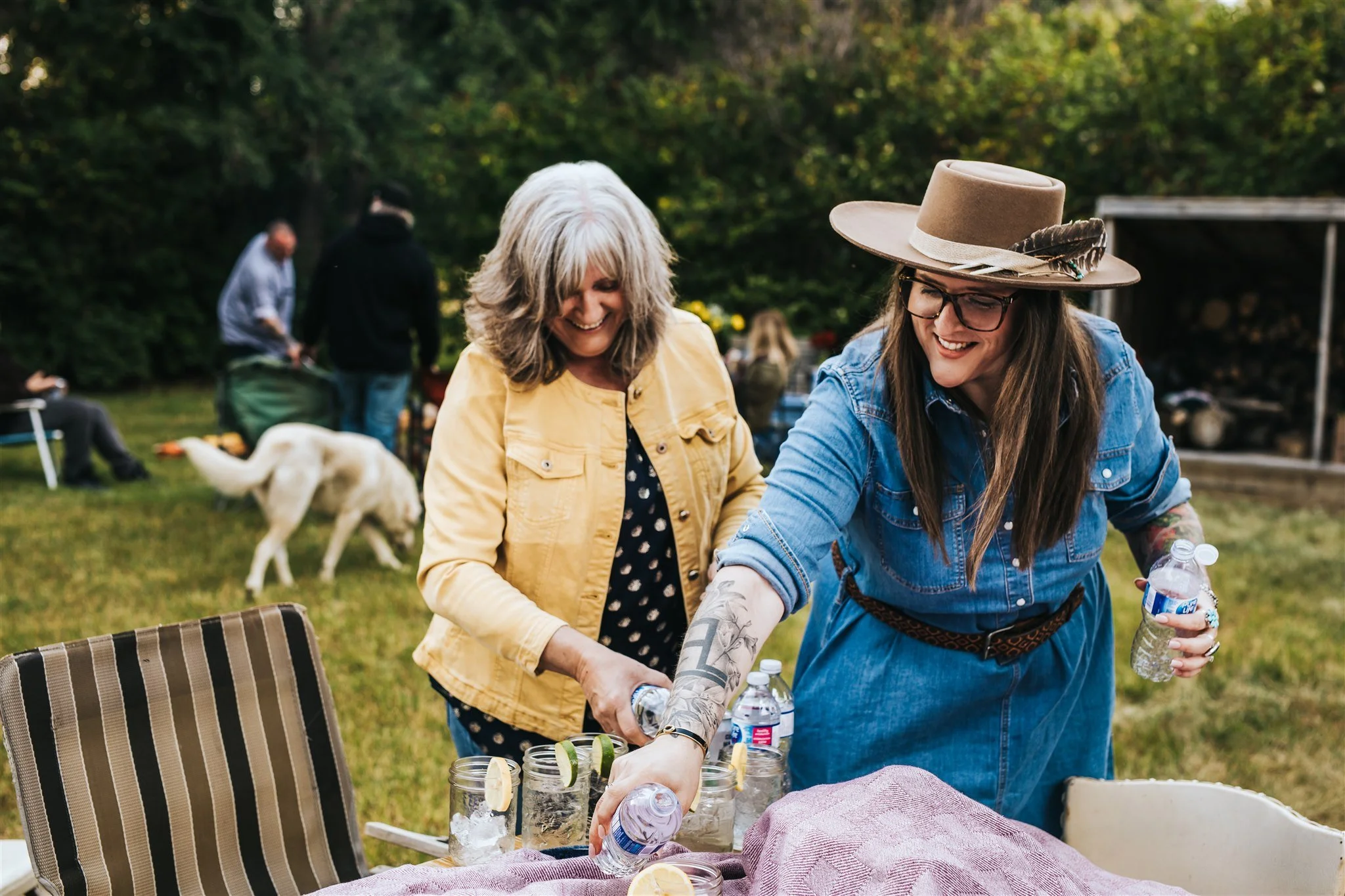 Two women at an outdoor gathering, smiling and pouring drinks into mason jars with lime slices, while a white dog walks in the background amidst trees and other people.