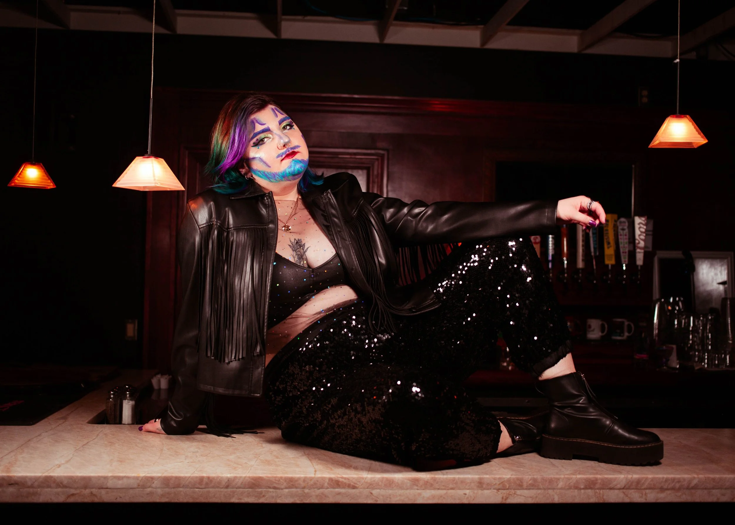 A person with colorful face paint, rainbow-colored hair, and multiple piercings poses in a dimly lit room with hanging lamps. They are wearing a black leather jacket, a sheer top with sparkles, and sequined black pants, sitting on a marble countertop.