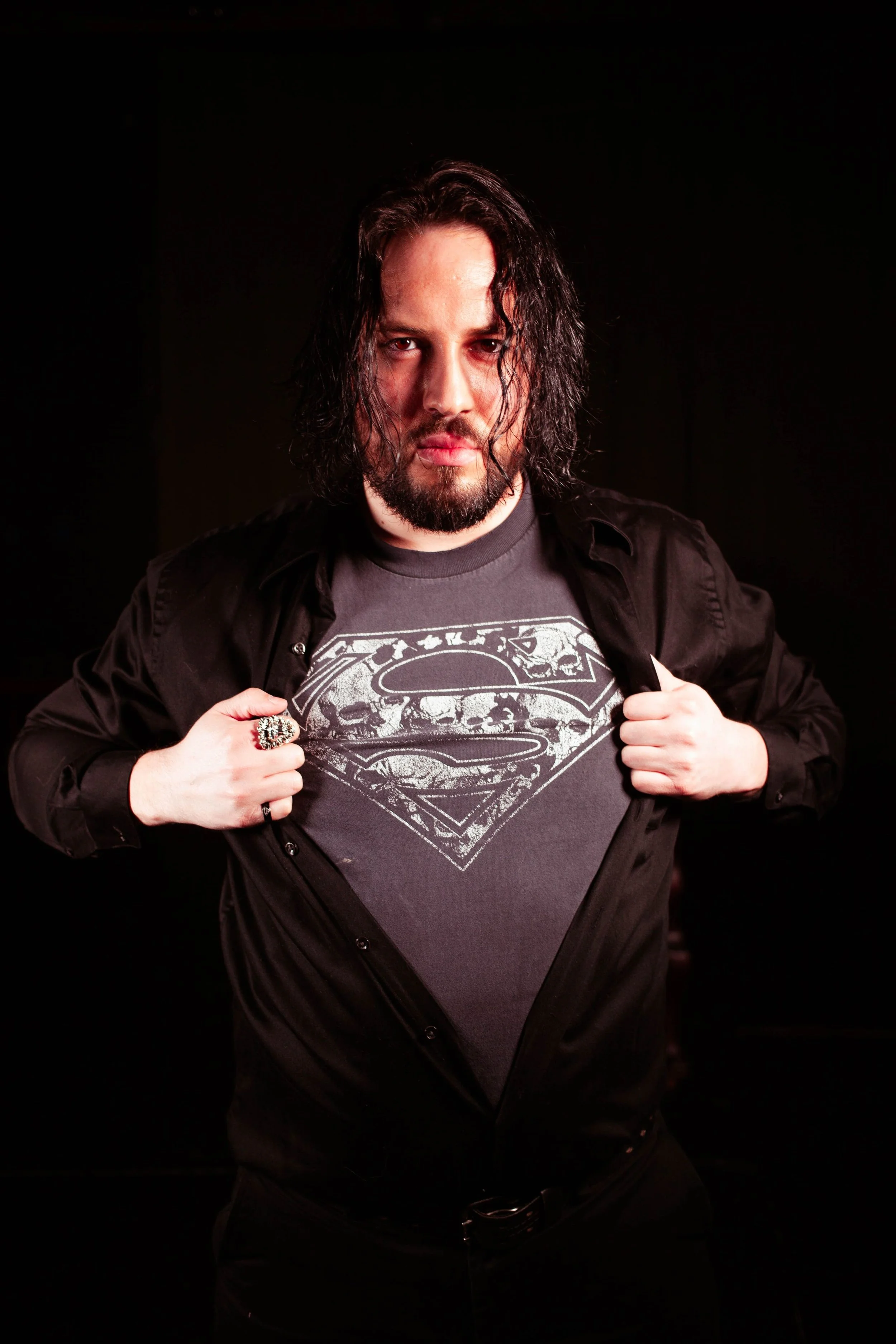 A man with long dark hair, beard, and mustache is wearing a black shirt over a gray T-shirt with a Superman logo. He is pulling open his shirt slightly with both hands, standing against a black background, with a serious expression.