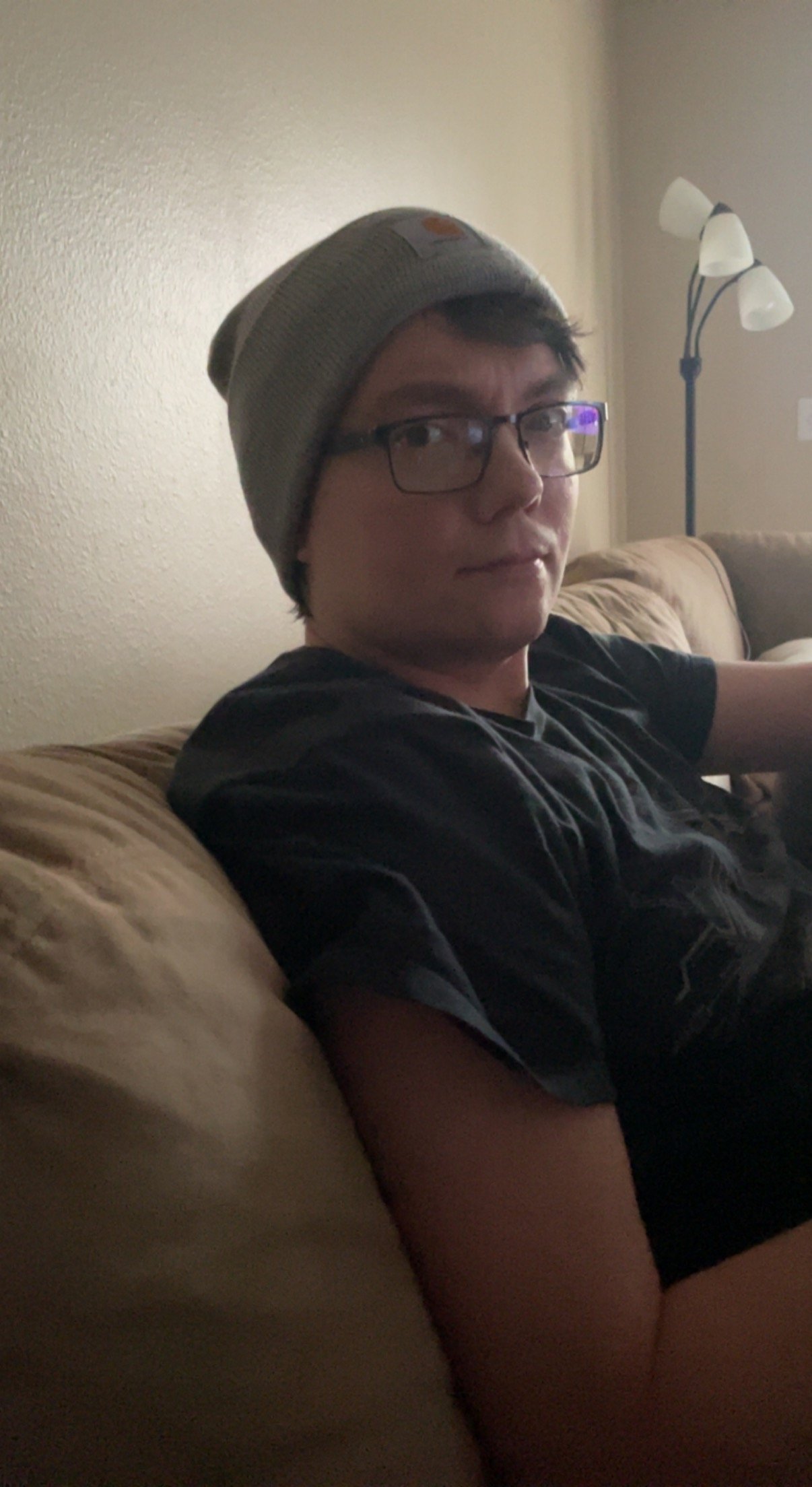 A young man with glasses wearing a gray beanie and a black t-shirt sitting on a beige couch in a living room, with a floor lamp in the background.