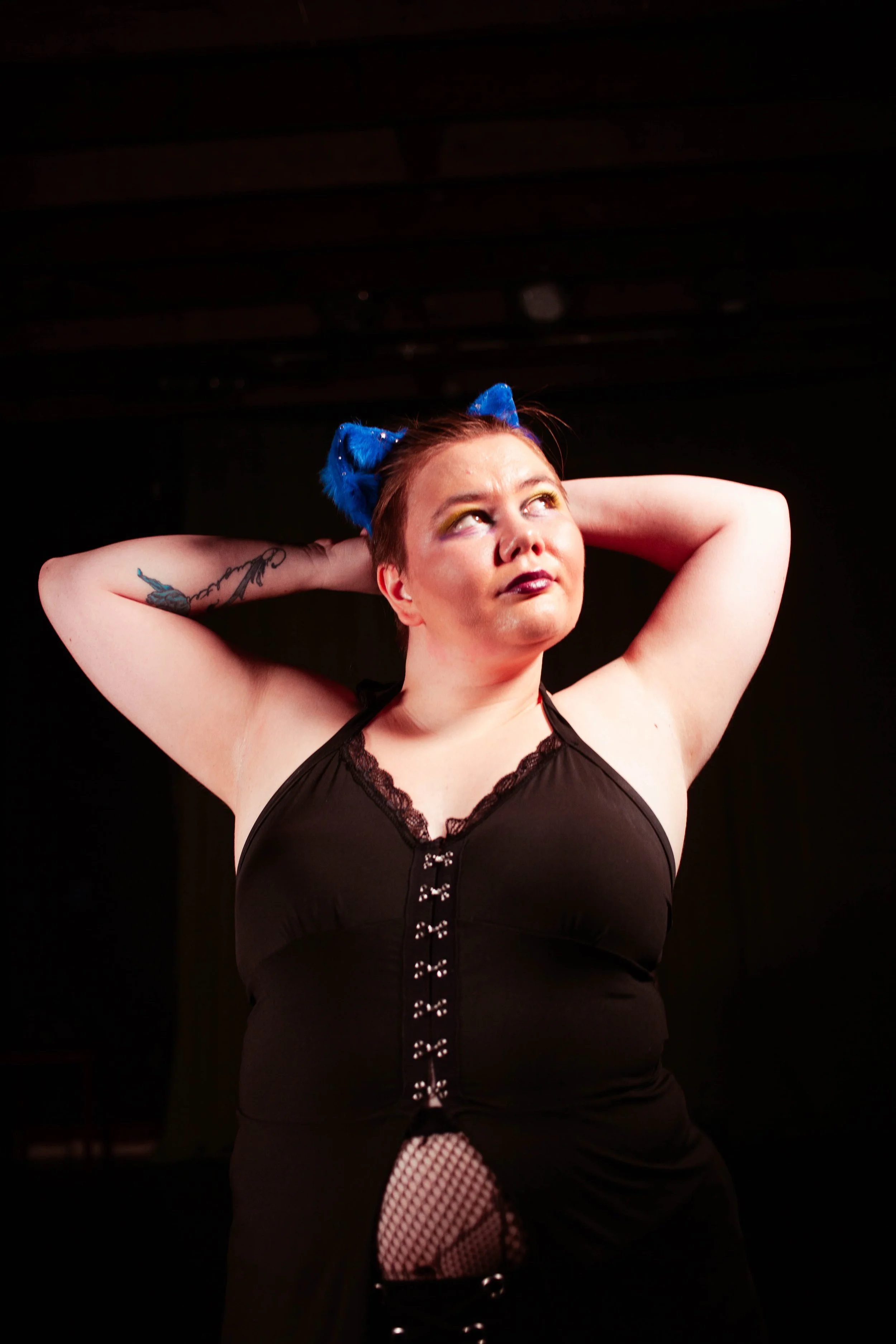 A person with short hair wearing a black top with hooked lace trim, blue fuzzy cat ears, yellow eyeshadow, and dark lipstick, posing with arms behind head against a black background.