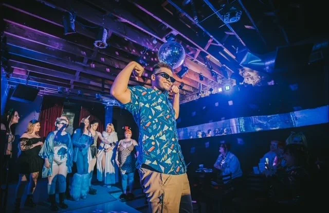 Man wearing sunglasses and a patterned shirt dancing or posing in a club or party setting with a disco ball overhead and a group of women dressed in costumes on stage in the background.