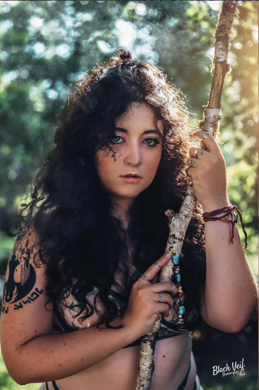 A young woman with dark, curly hair and tattoos on her arm, holding a stick decorated with beads, in a forest setting with sunlight filtering through the trees.