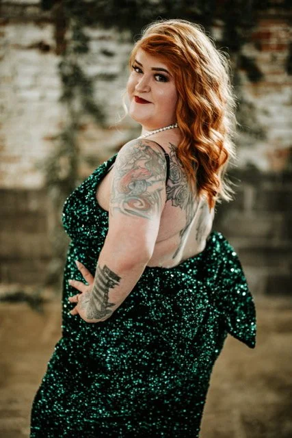 Red-haired woman with tattoos wearing a sparkly green dress, posing outdoors with a brick wall background.