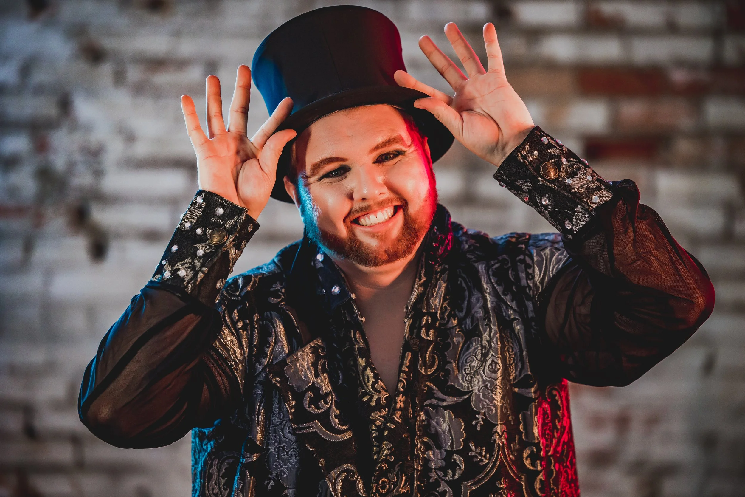 A man with makeup and a beard, wearing a patterned jacket and a black top hat, posing with hands near the hat against a blurred background.
