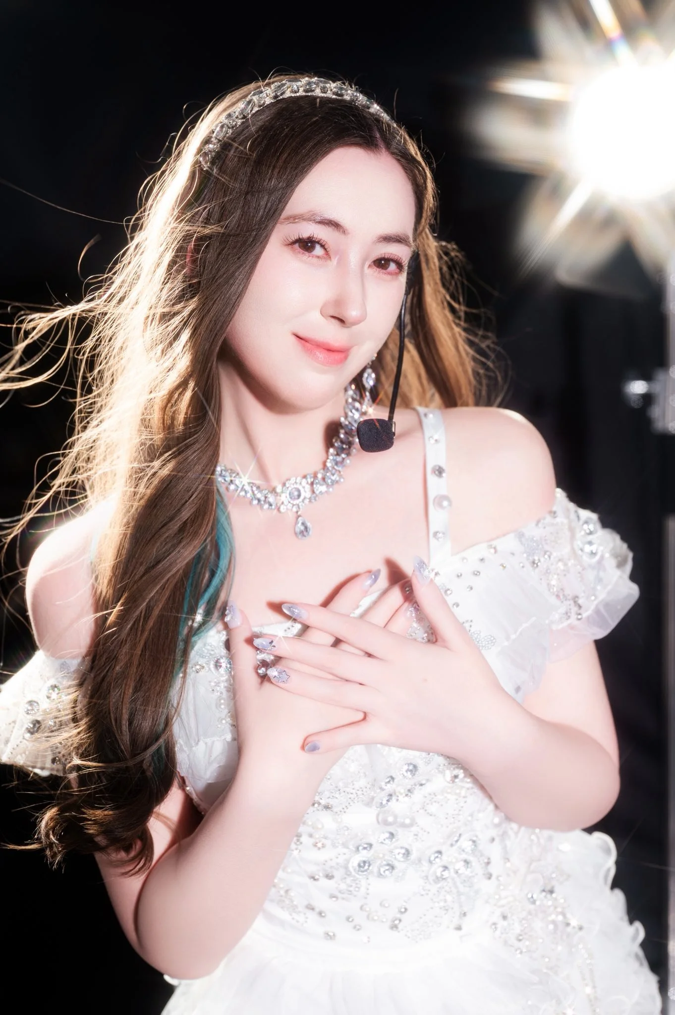 A woman with long brown hair and a pearl headband wearing a white dress with rhinestones, smiling softly with her hands crossed over her chest. She wears a necklace and earrings, with a light source glowing behind her on the right side.
