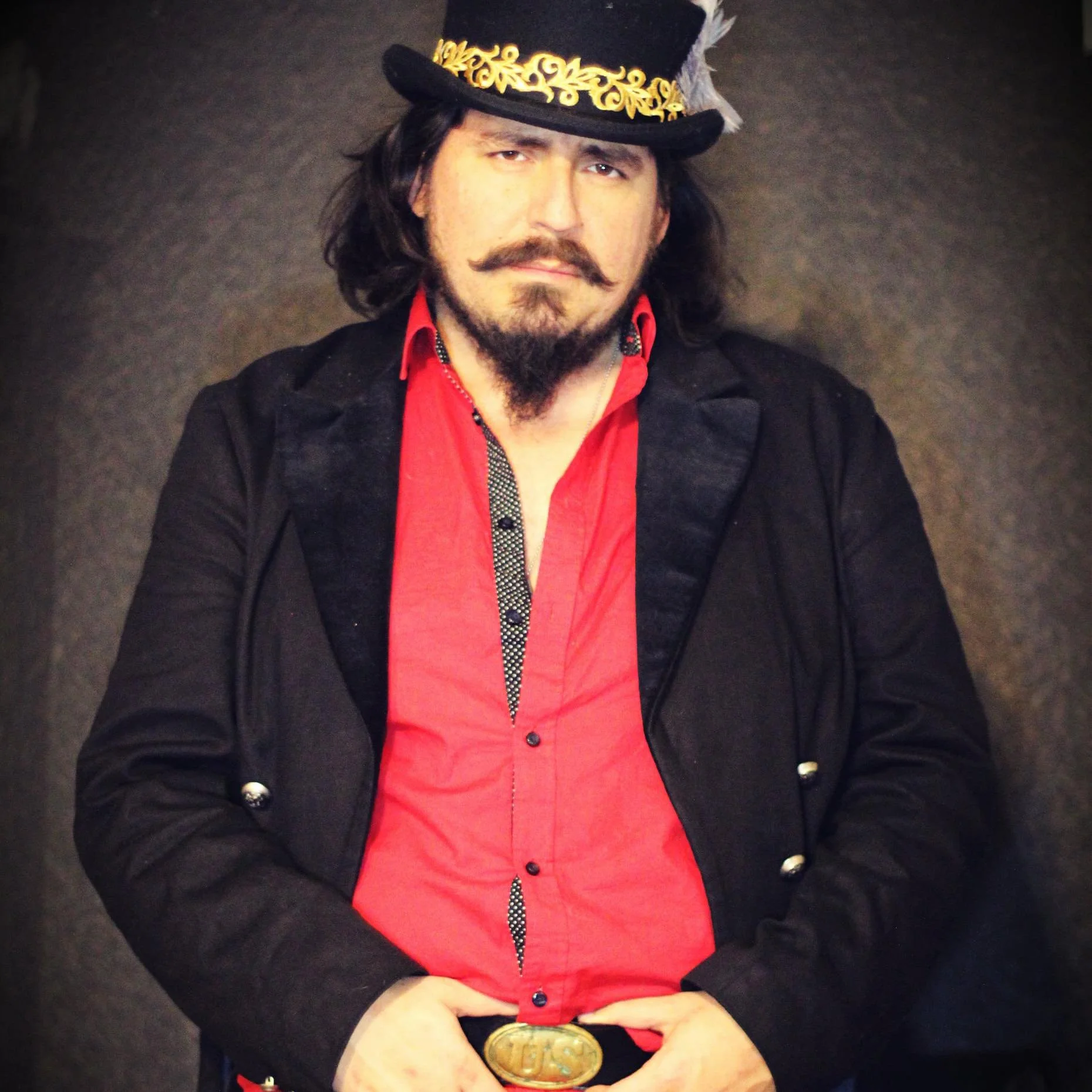 Man in a black jacket, red shirt, gold belt buckle, and a black top hat with gold embroidery, standing against a dark background.