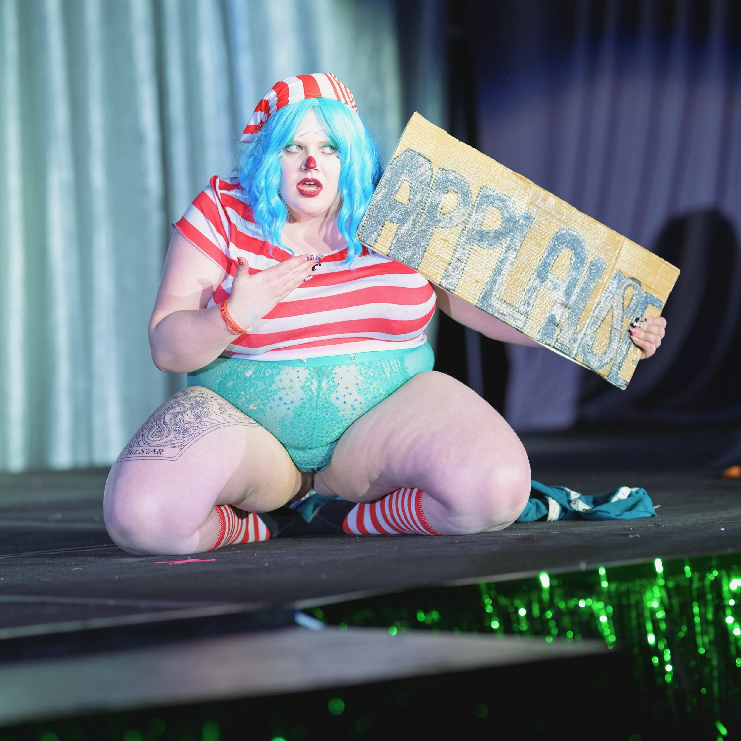 A performer dressed as Buggy the Clown with blue hair, red nose, makeup, striped red and white shirt, plaid cap, holding a glittery Applause sign on a stage.
