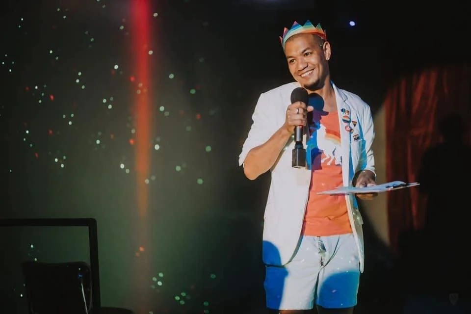 A man wearing a paper crown, white blazer, and orange shirt, smiling and holding a microphone and papers on a stage with a dark background and colorful lights.