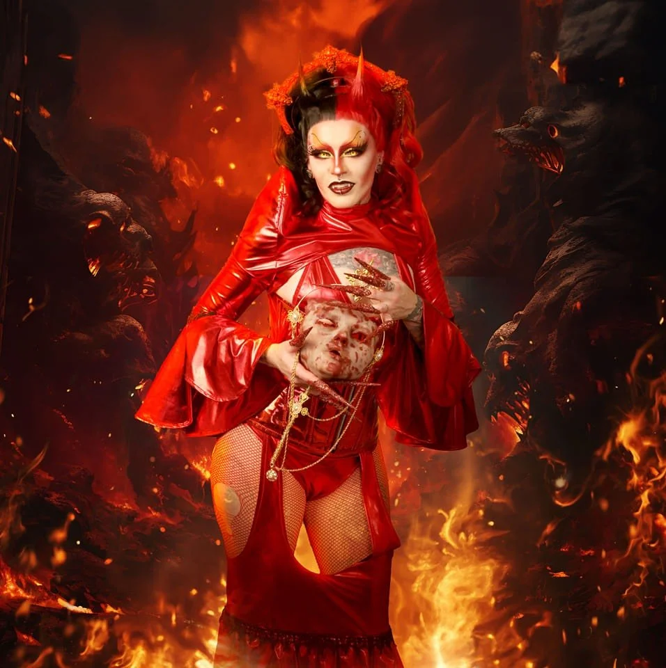 A person dressed as a devil with red hair, horns, and red leather clothing, holding a severed head prop in a fiery, hellish background with demonic faces.