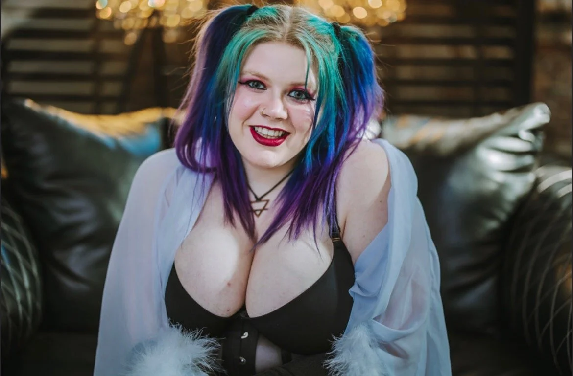 A woman with colorful rainbow hair and a black bra, smiling at the camera while sitting on a black leather couch.