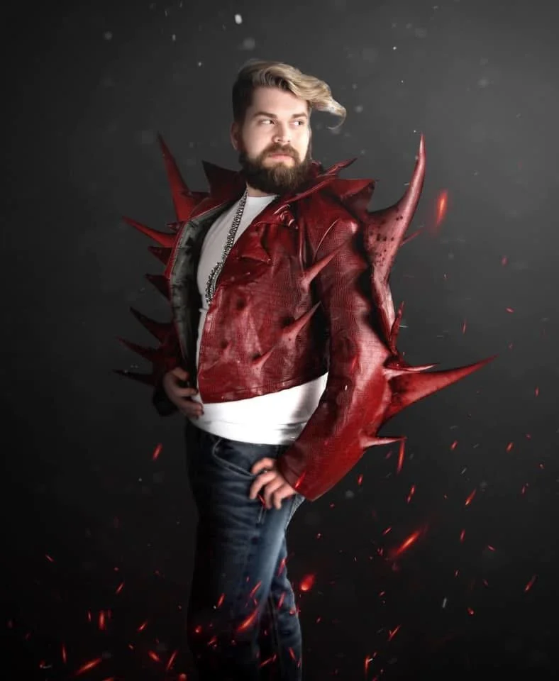 A person with a beard and styled hair in a red, spiked, dragon-shaped jacket posing against a dark background with red sparks.