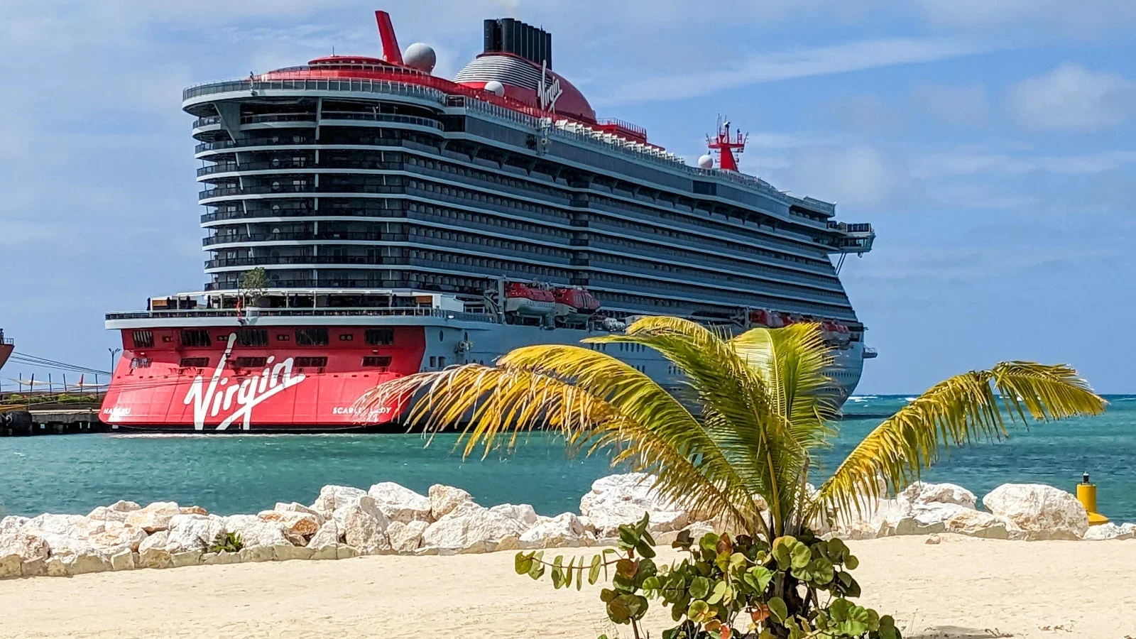A large cruise ship named Virgin docked at a tropical port, with palm trees and rocks in the foreground, and clear blue sky and ocean in the background.