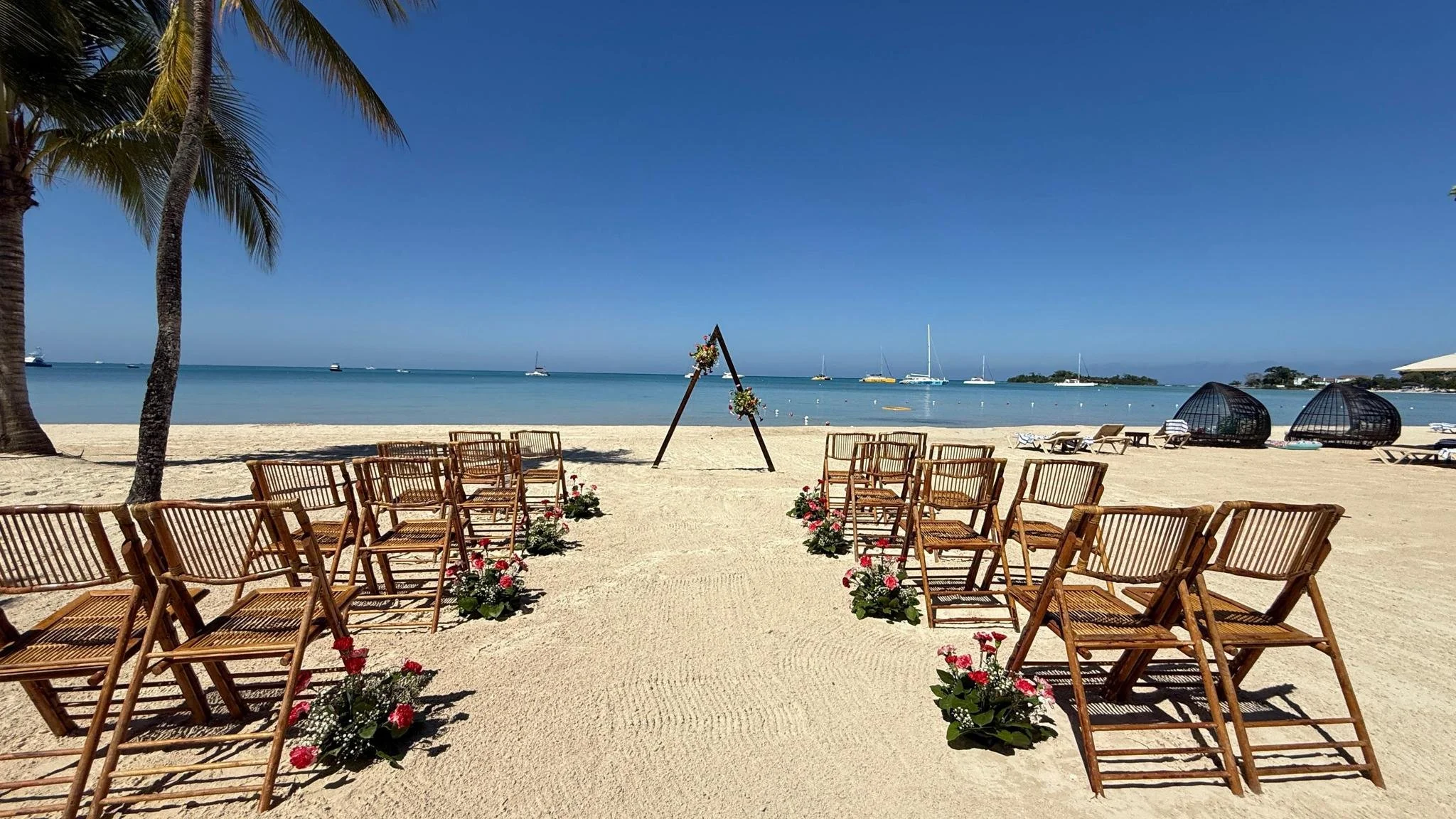 A beach setup for a wedding ceremony with wooden chairs arranged on the sandy shore, decorated with flowers, facing the ocean. There are lounge chairs, two dome-shaped cabanas, a few boats on the water, and a clear blue sky at Sandals Resorts