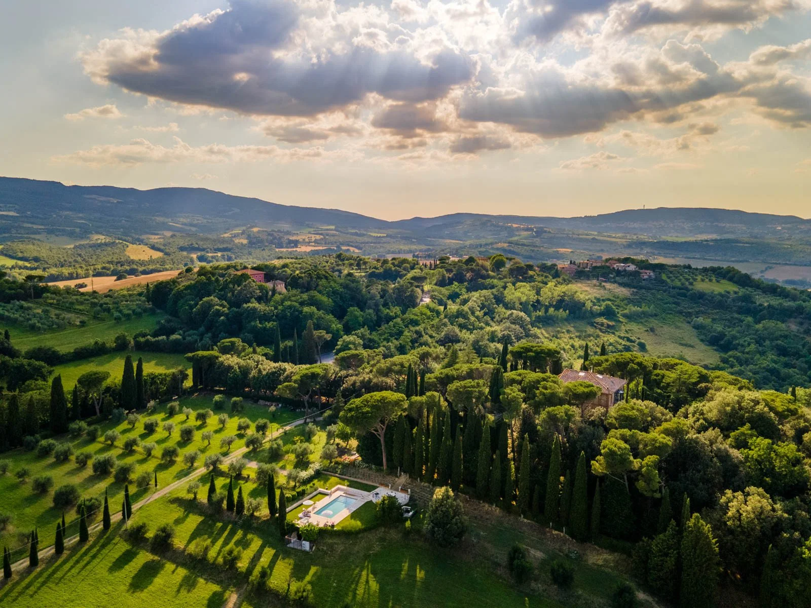 A scenic landscape of rolling green hills, tall cypress trees, and scattered houses under a partly cloudy sky.