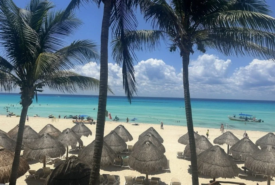 A beach scene featuring palm trees, thatched umbrellas, lounge chairs, and people enjoying the sunny weather by turquoise ocean water with a boat in the background.