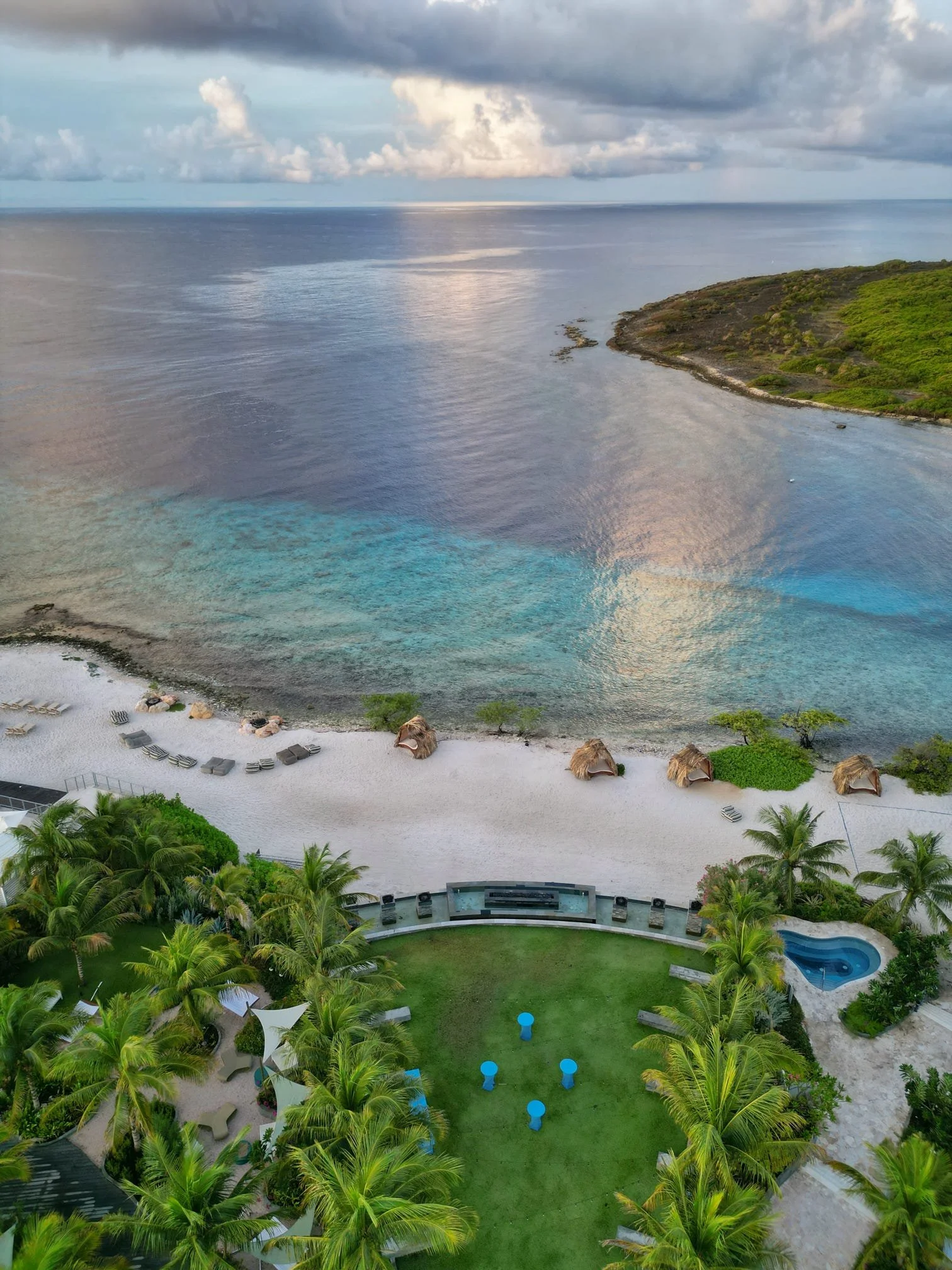 Aerial view of a tropical resort with green palm trees, a grassy area with blue stools, a small pool, sandy beach with huts and lounge chairs, and calm turquoise water meeting the horizon under cloudy skies.