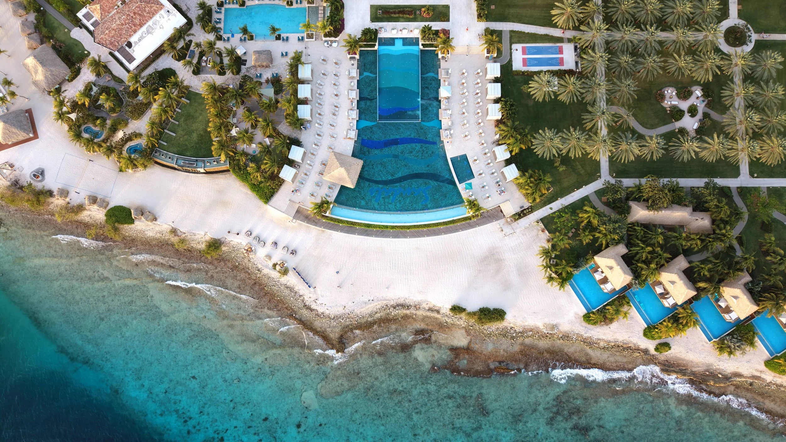 Aerial view of a luxurious beachfront resort with swimming pools, lounge chairs, palm trees, and cabanas along a sandy beach with ocean waves.