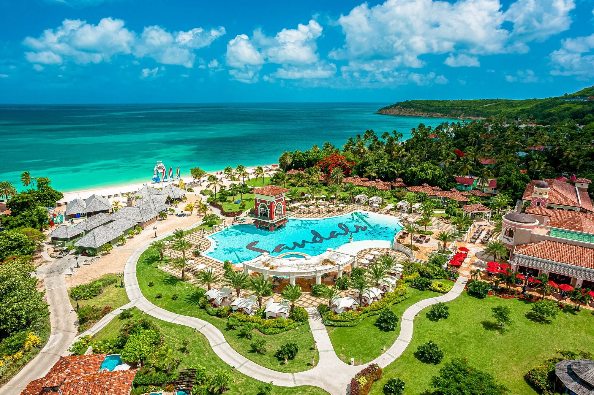 Aerial view of a tropical resort featuring a large swimming pool with the word 'Sandals' written in it, surrounded by palm trees, umbrellas, and lush greenery. The resort overlooks a turquoise ocean with a sandy beach and small huts, with a backdrop of green hills and a partly cloudy sky.