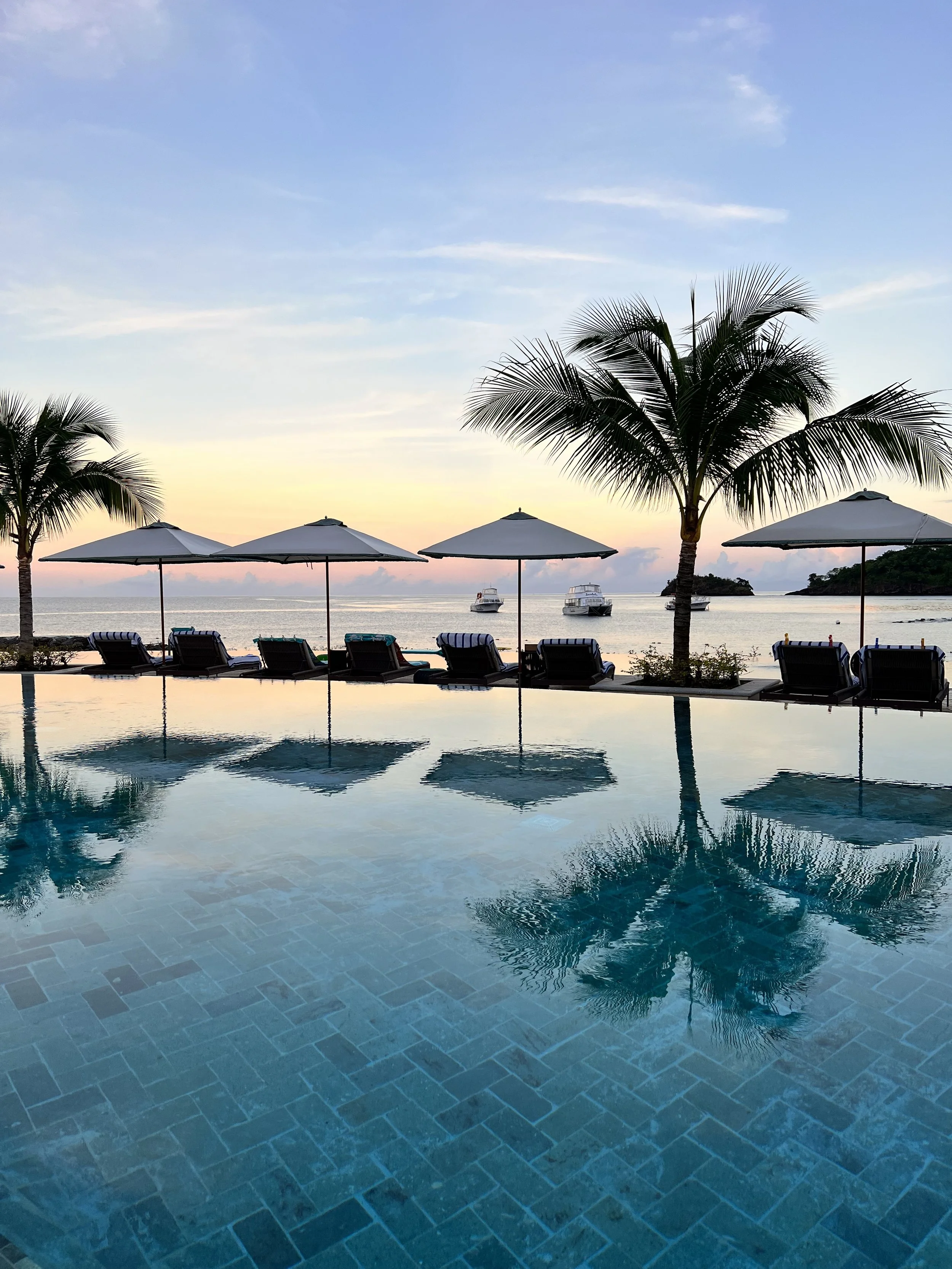 A tropical beach scene at sunset with a pool in the foreground, empty lounge chairs, large white umbrellas, palm trees, boats on the water, and a small island in the distance under a partly cloudy sky.
