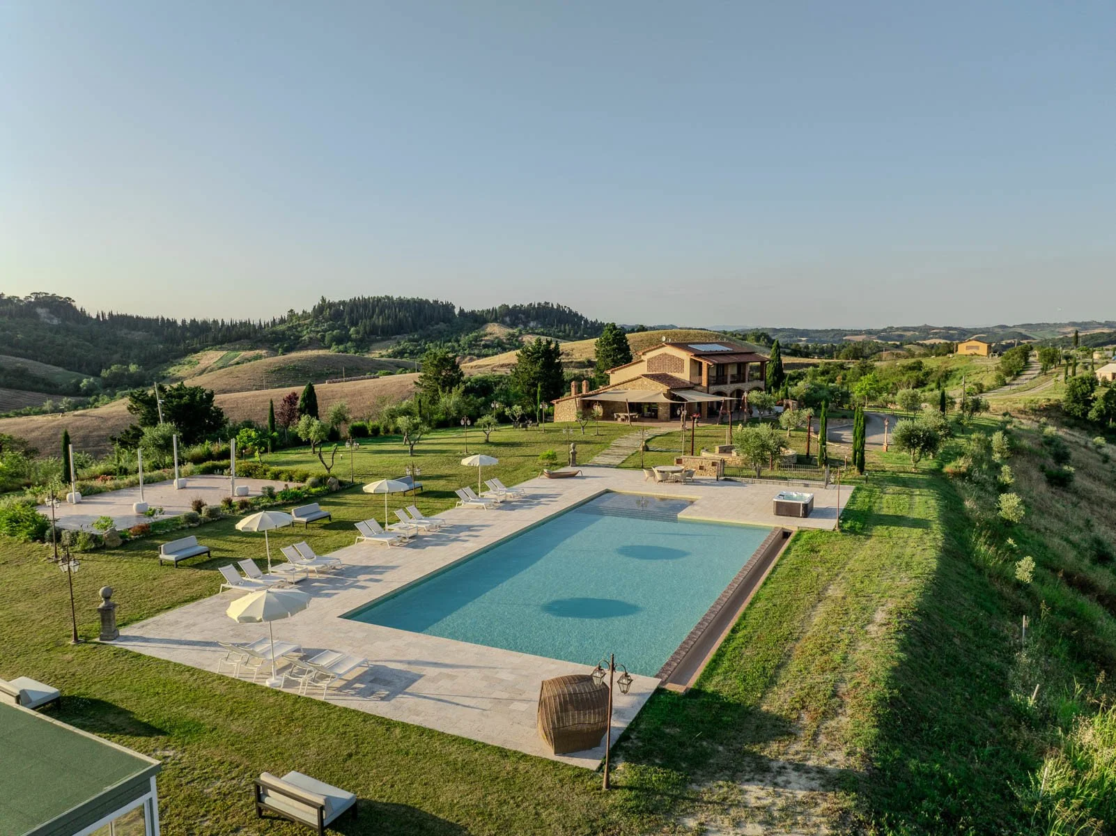 A scenic view of a luxurious house with a swimming pool, surrounded by green lawns, umbrellas, lounge chairs, and scenic hills in the background.