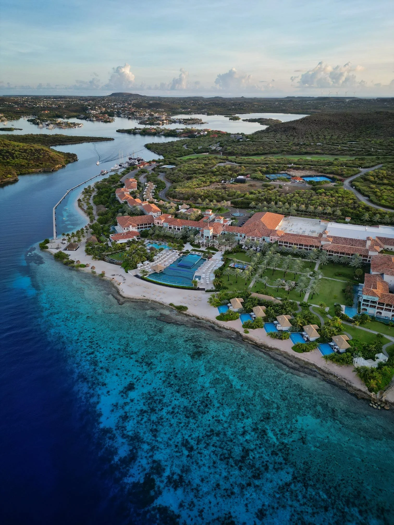 Aerial view of a luxury resort on a coastline with beachfront pools, cabanas, palm trees, and a large building with orange rooftops, surrounded by green hills and a body of water.