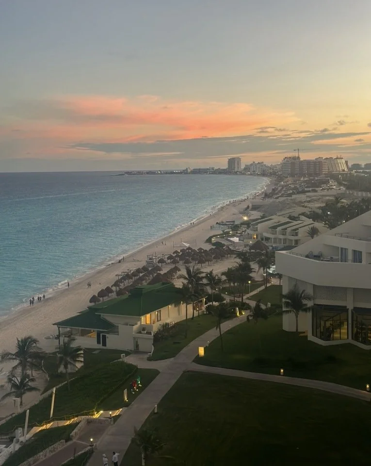 A sunset view of a beach with people walking along the shoreline, modern beachfront buildings, palm trees, and a city skyline in the distance.