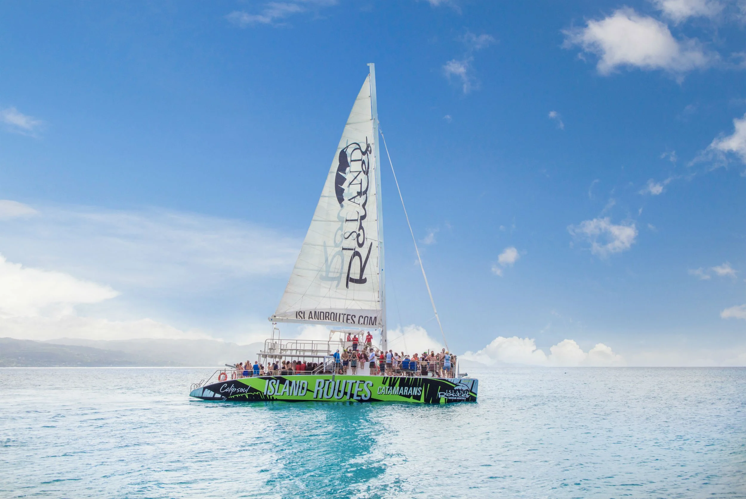 A catamaran boat named 'Island Routes' is sailing on a calm ocean with a large crowd of people on board. The boat has a tall sail with the website 'islandroutes.com' and the word 'catamarans' written on it, and the sky above is mostly clear with some scattered clouds.