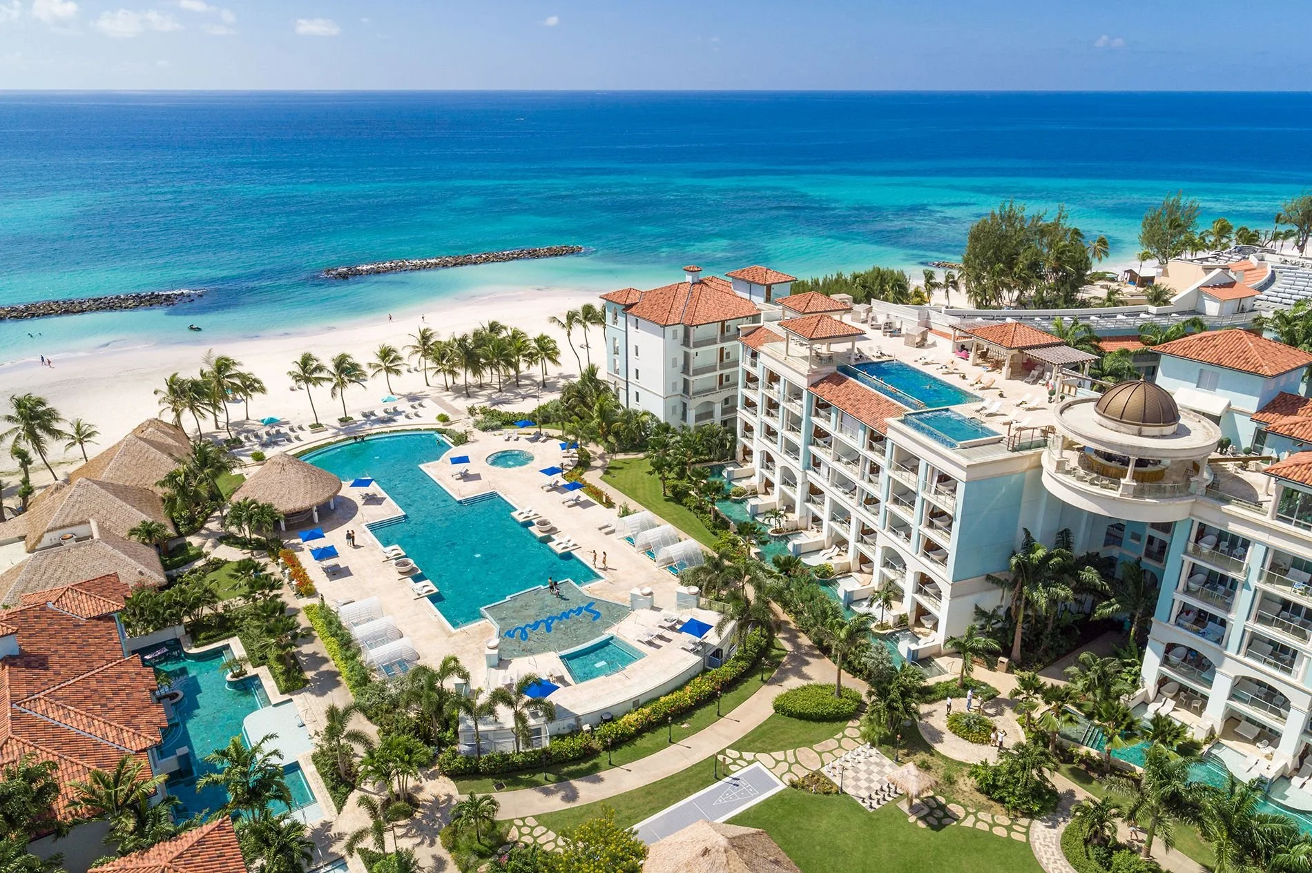 High-angle aerial view of a beachfront resort with multiple swimming pools, palm trees, lounge chairs, and buildings with red-tiled roofs, overlooking a sandy beach and the blue ocean.