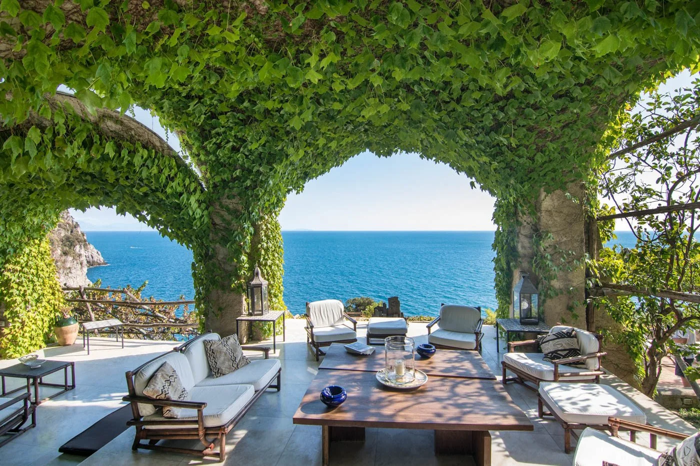 Outdoor seating area with white cushioned chairs and sofas under a arched vine-covered structure overlooking the ocean.