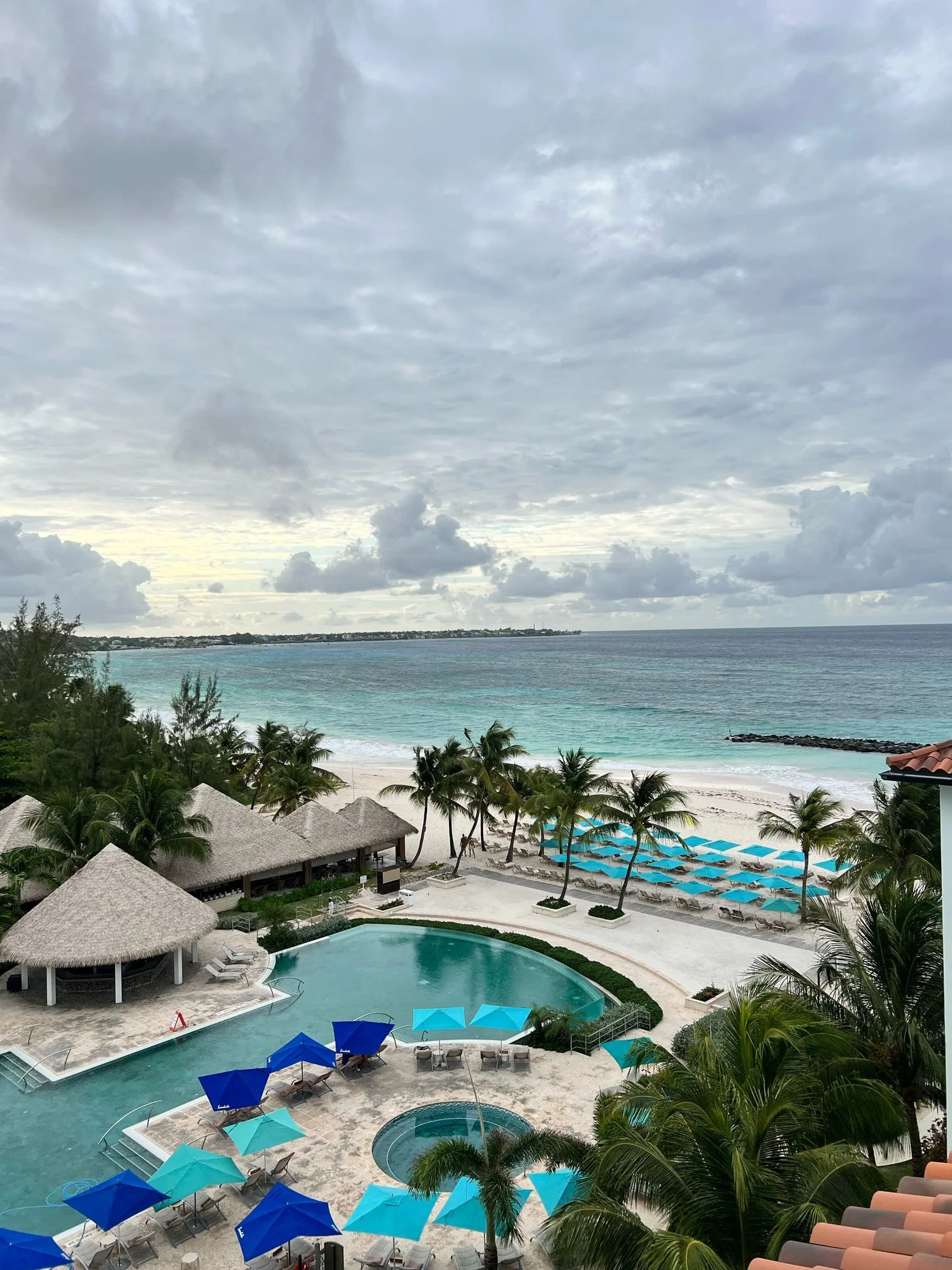 View of a tropical resort with a swimming pool, lounge chairs with blue umbrellas, thatched roof structures, palm trees, and the ocean in the background under a cloudy sky.