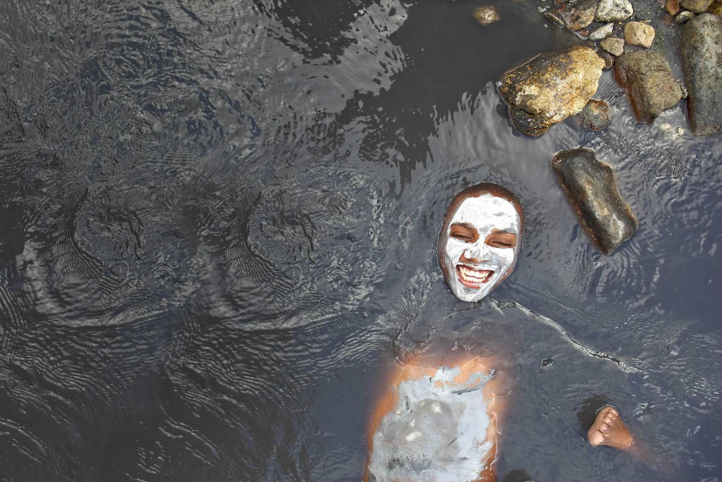 A woman with white face paint and a big smile, floating in dark water with rocks around her, her head and one foot visible.