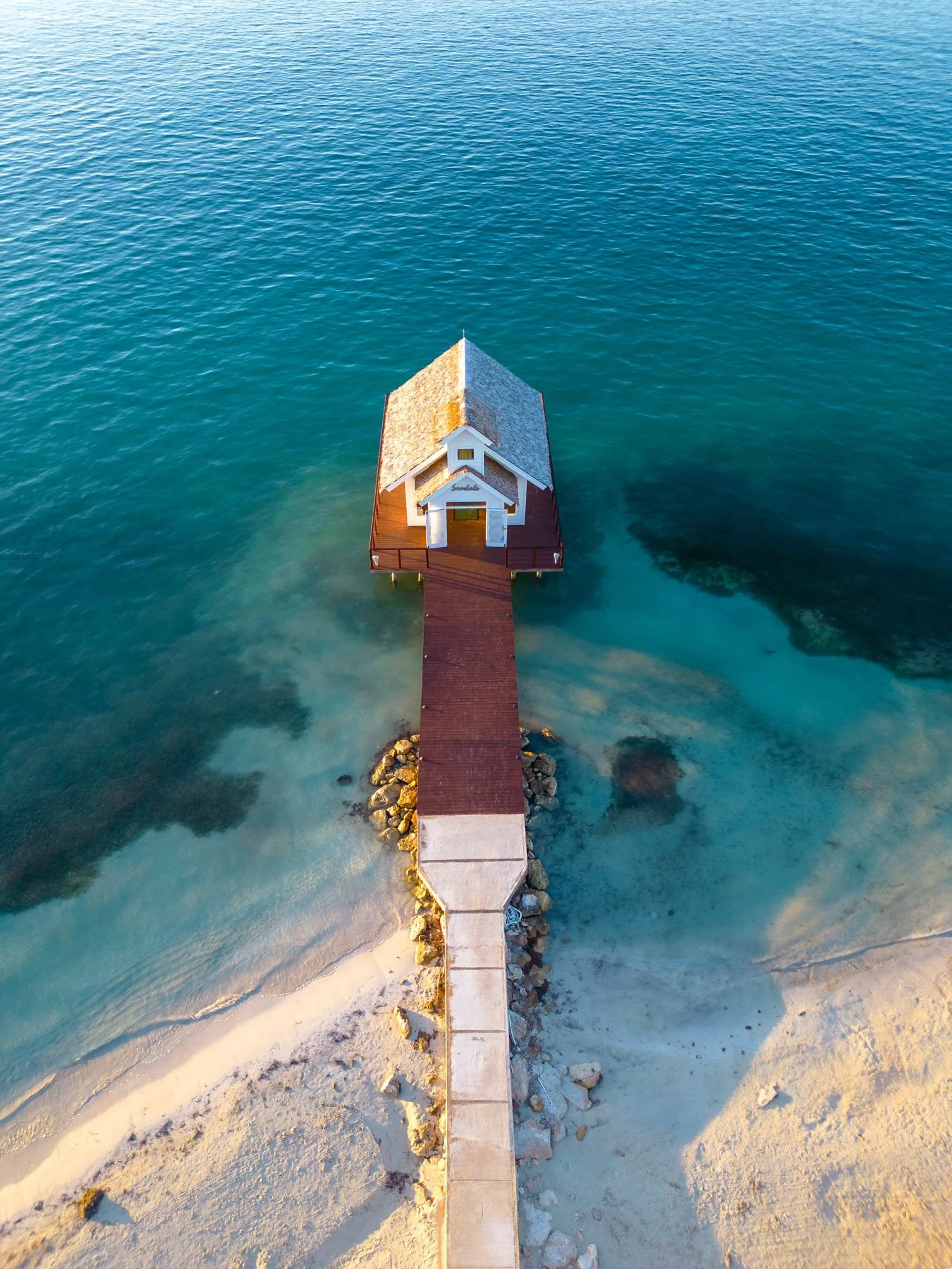 An aerial view of a small pier leading to a house on stilts over the water, with clear blue and green ocean surrounding it and sandy beach at the shoreline.