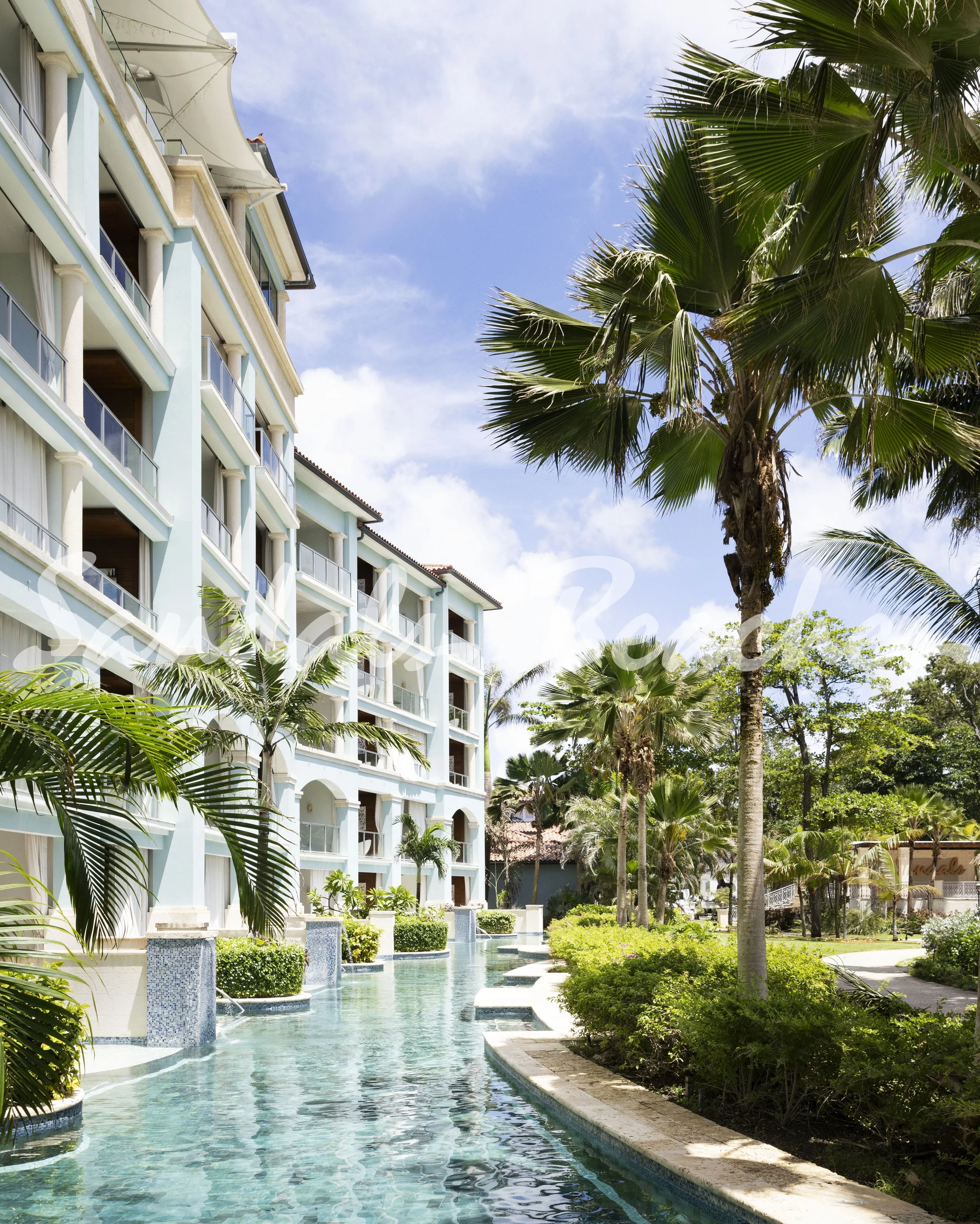 A resort with a swimming pool surrounded by tropical palm trees, with a multi-story building in the background, under a blue sky with some clouds.