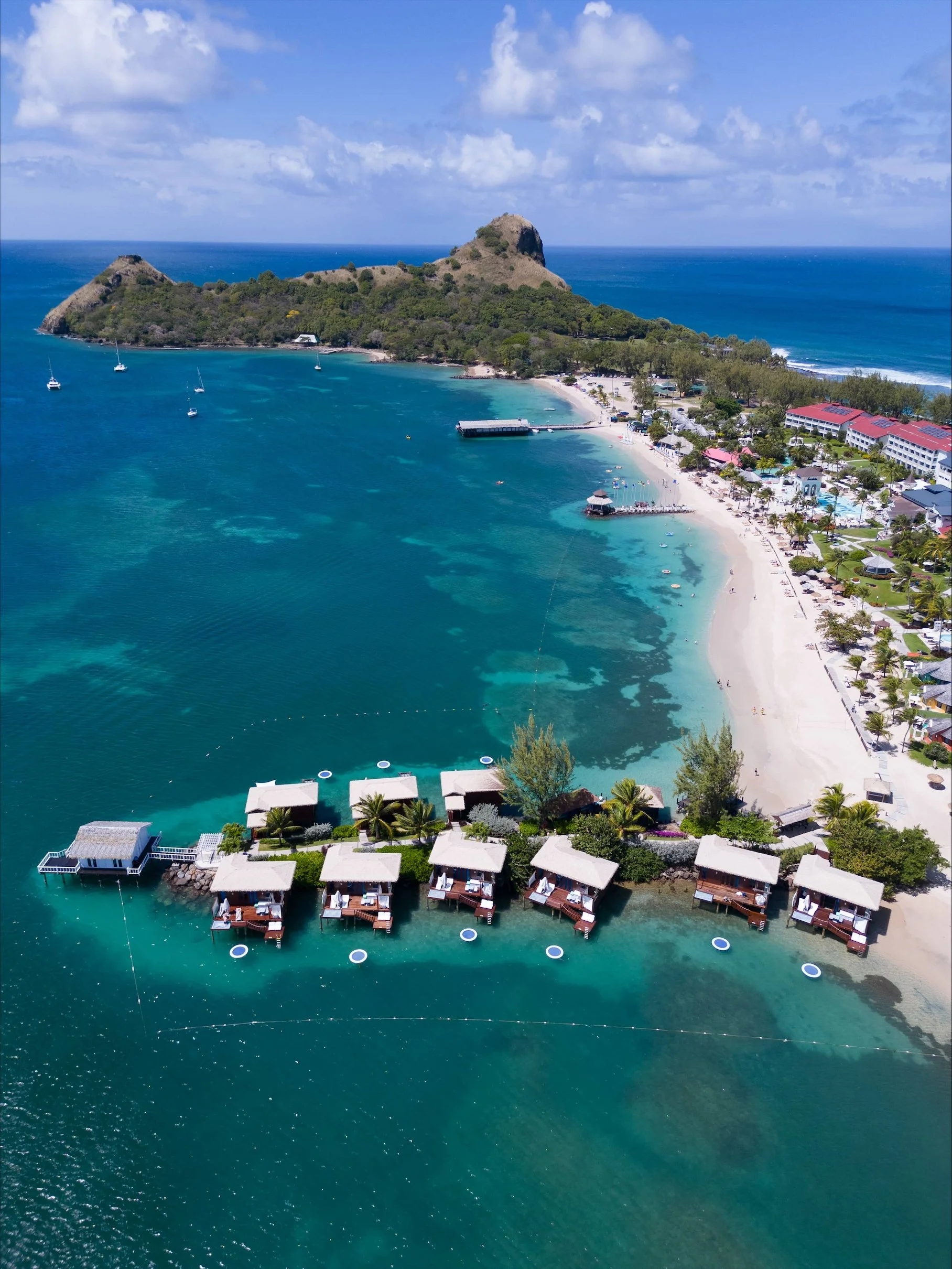 Aerial view of a tropical beach with overwater bungalows, a sandy shoreline, boats in the turquoise water, and a green island with a mountain in the background.