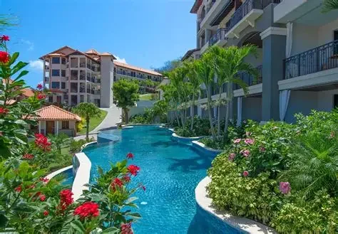 A luxurious swimming pool surrounded by lush green plants and flowers, with multi-story residential buildings in the background under a clear blue sky.