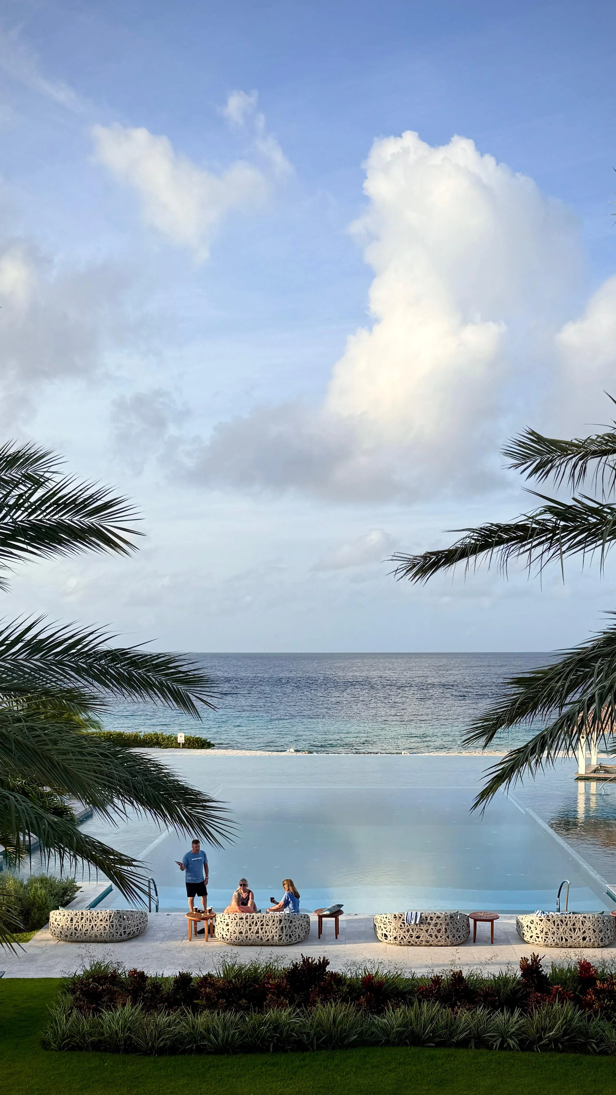 People relaxing by a pool with a view of the ocean and sky, framed by palm trees.