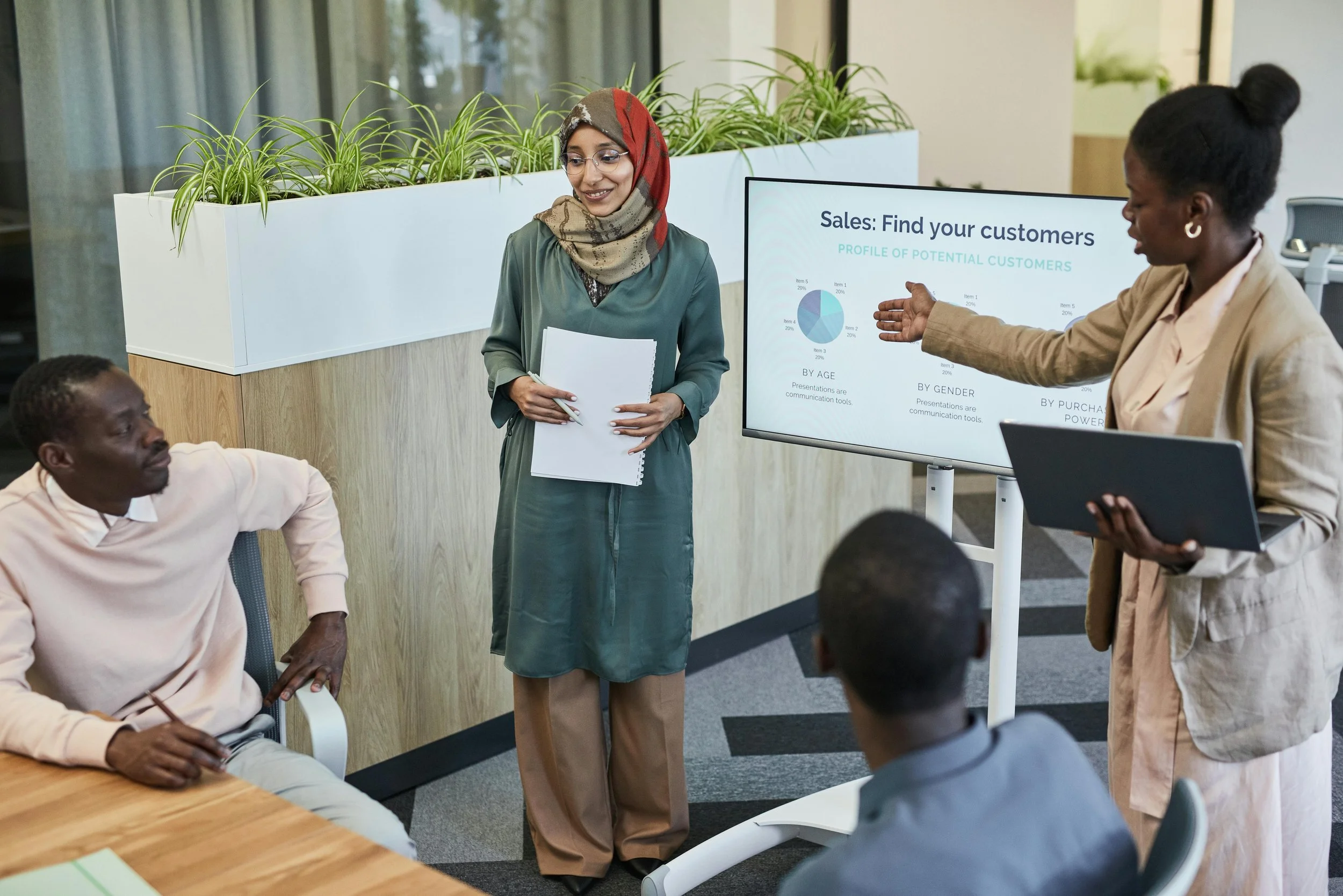 A diverse group of people attending a presentation in an office, with a woman standing by a screen showing sales data and charts, and others seated around a table listening.