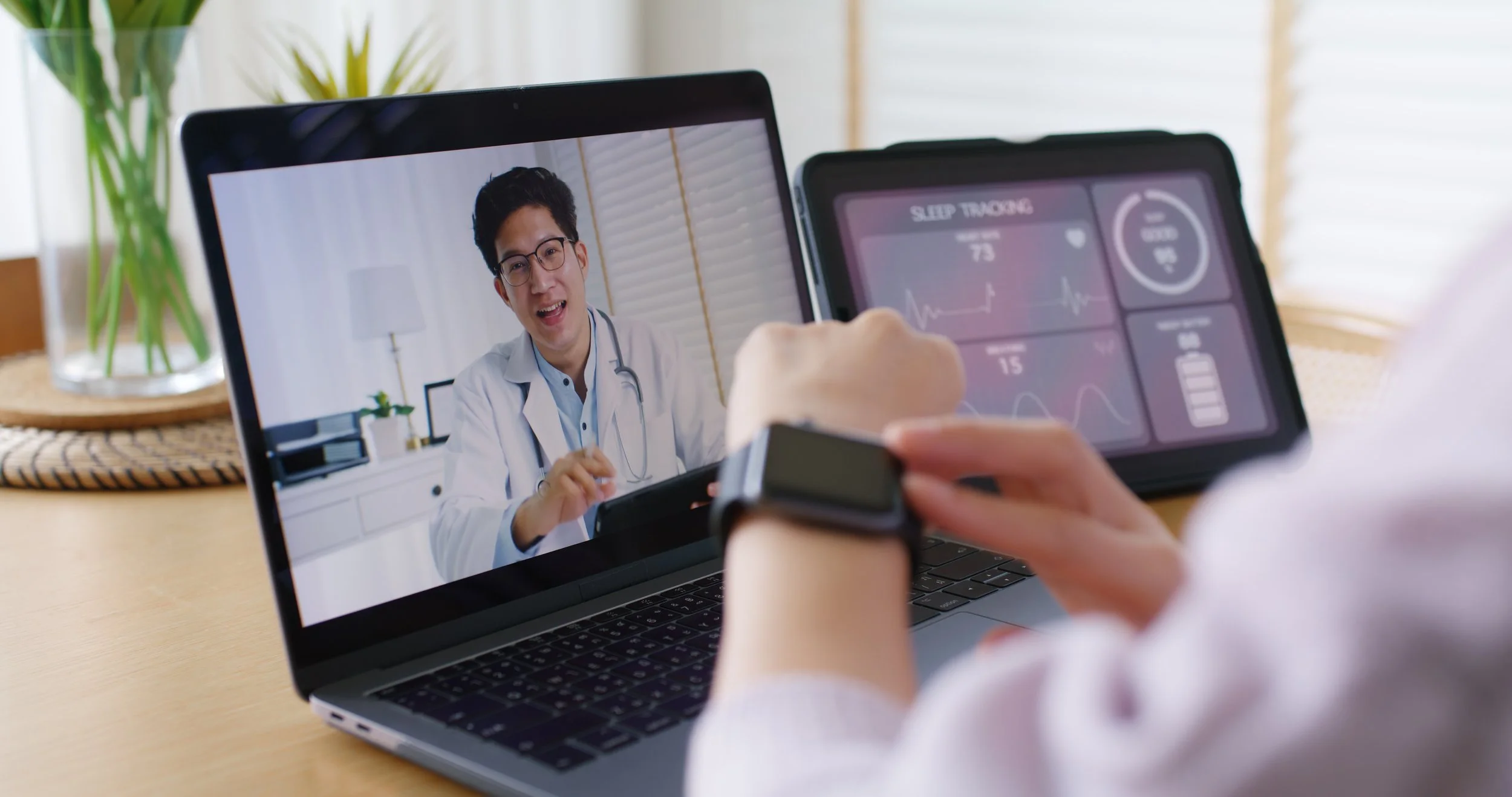 A healthcare professional, wearing blue scrubs, is taking notes on a notepad while holding a tablet displaying reading material. A stethoscope is on the white desk next to the tablet, and sunglasses are nearby.