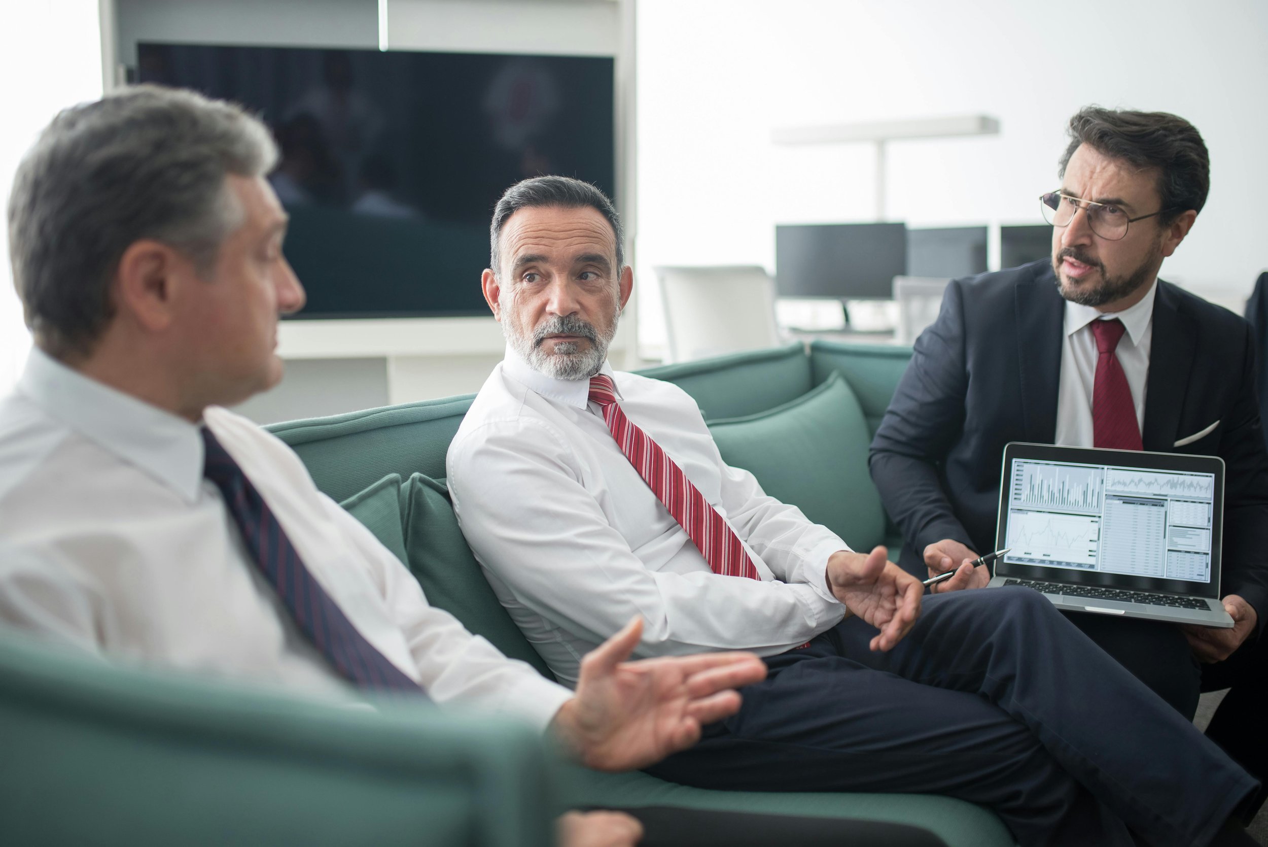 Three business professionals in formal attire having a discussion in an office conference room, with one holding a laptop displaying charts and graphs.